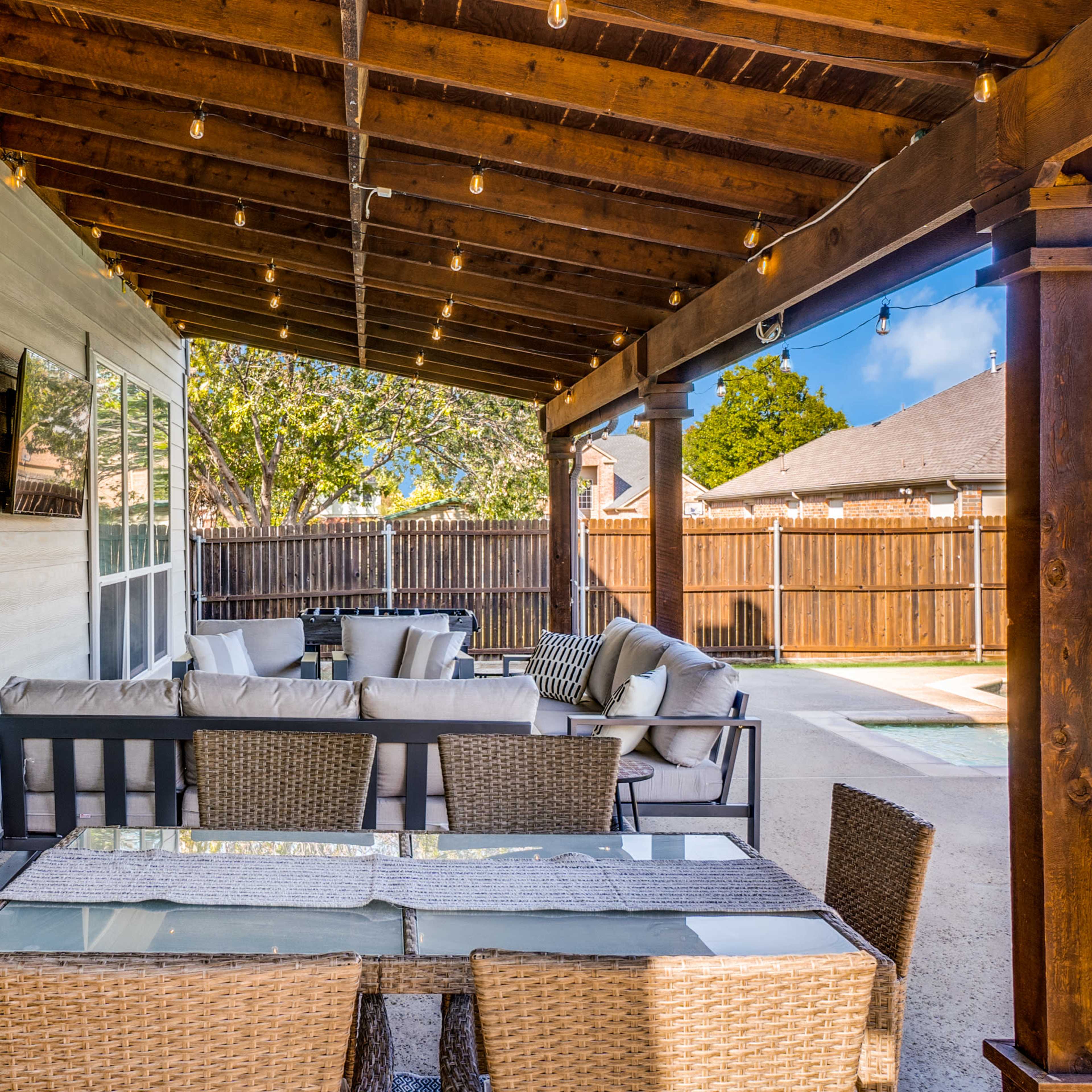 A covered outdoor patio with a dining table and seating area, overlooking a swimming pool and surrounding fenced yard.