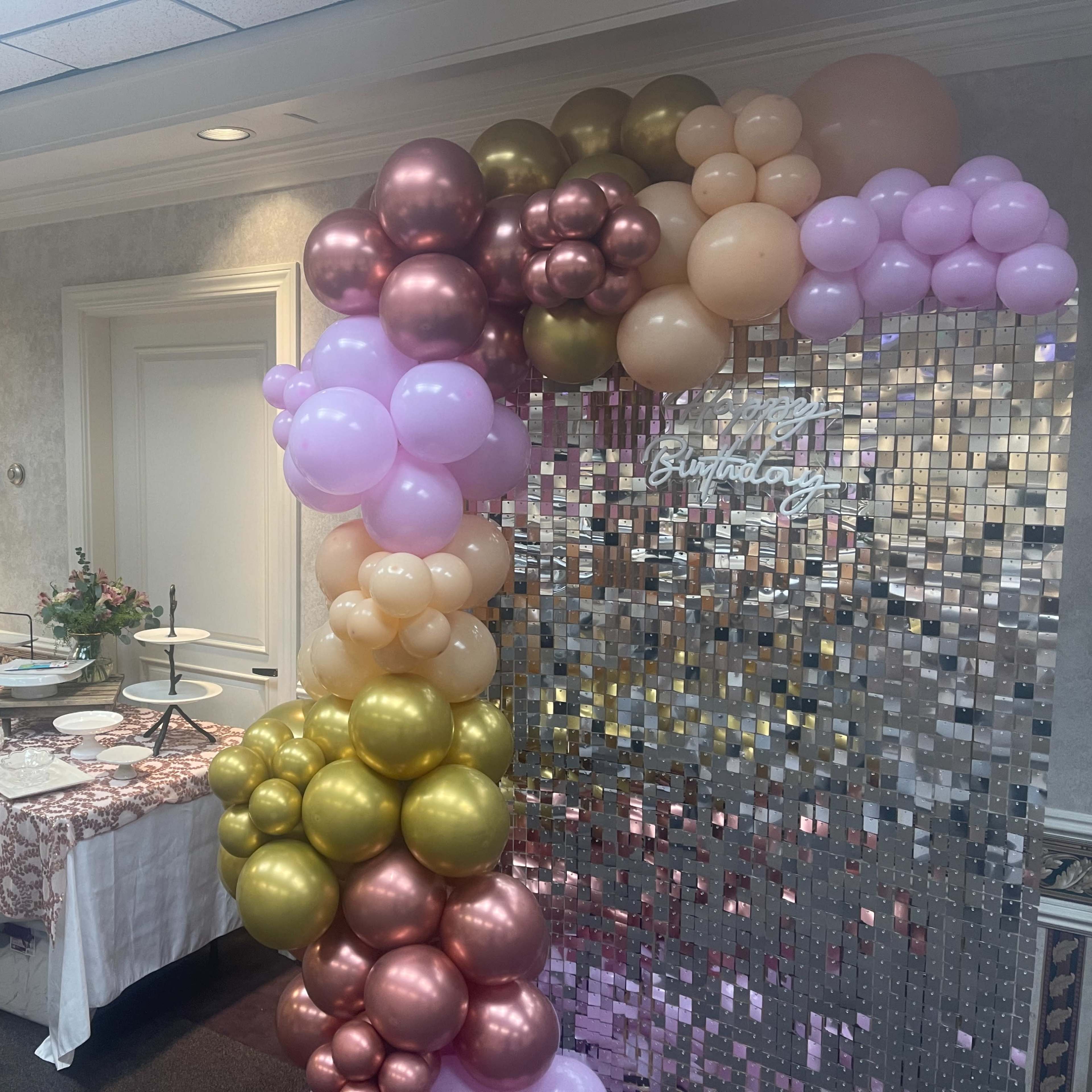A table with desserts is set up next to a balloon arch and a shiny backdrop that says "Birthday."