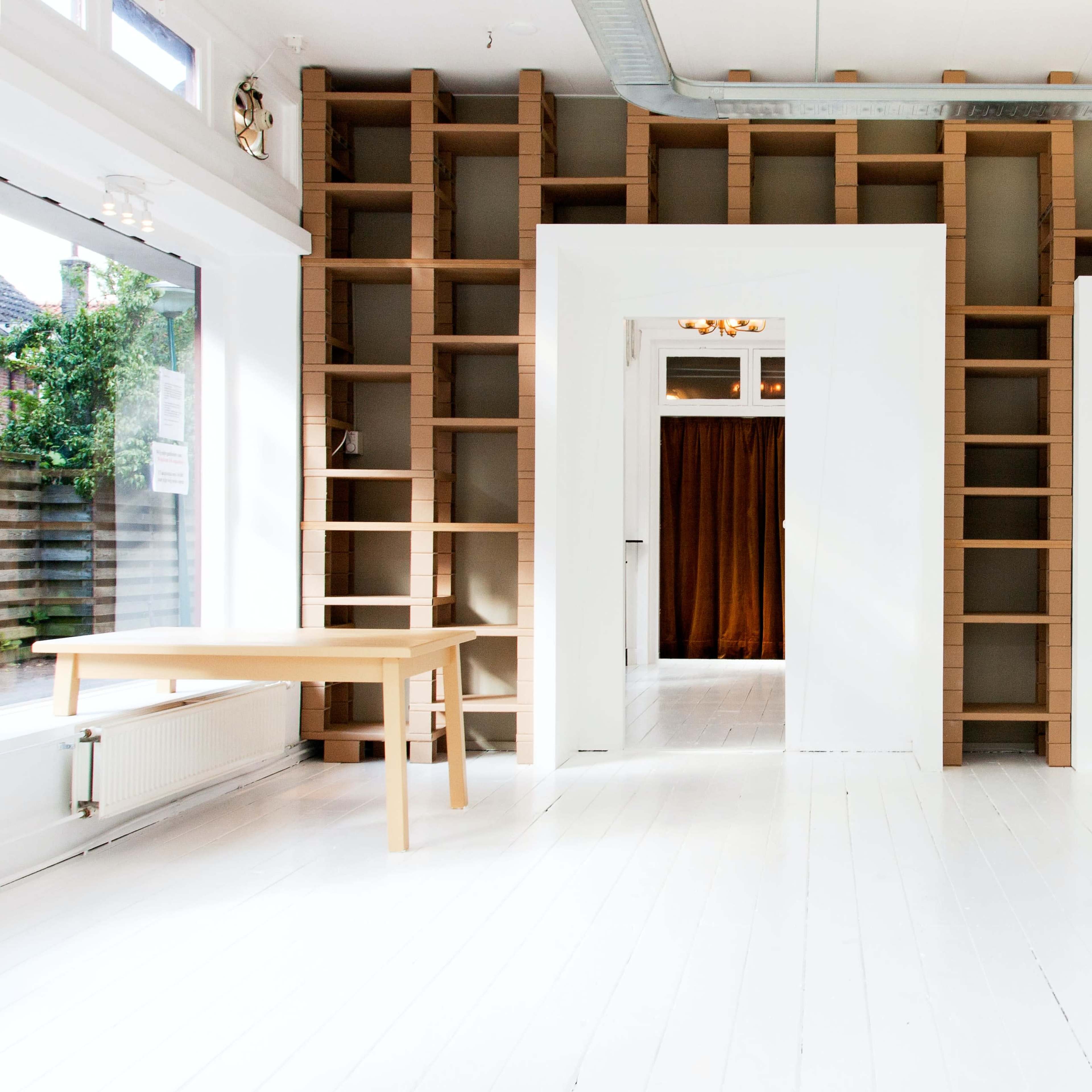 A brightly lit interior space with white floors, minimalist wooden furniture, and a wall featuring open shelving.