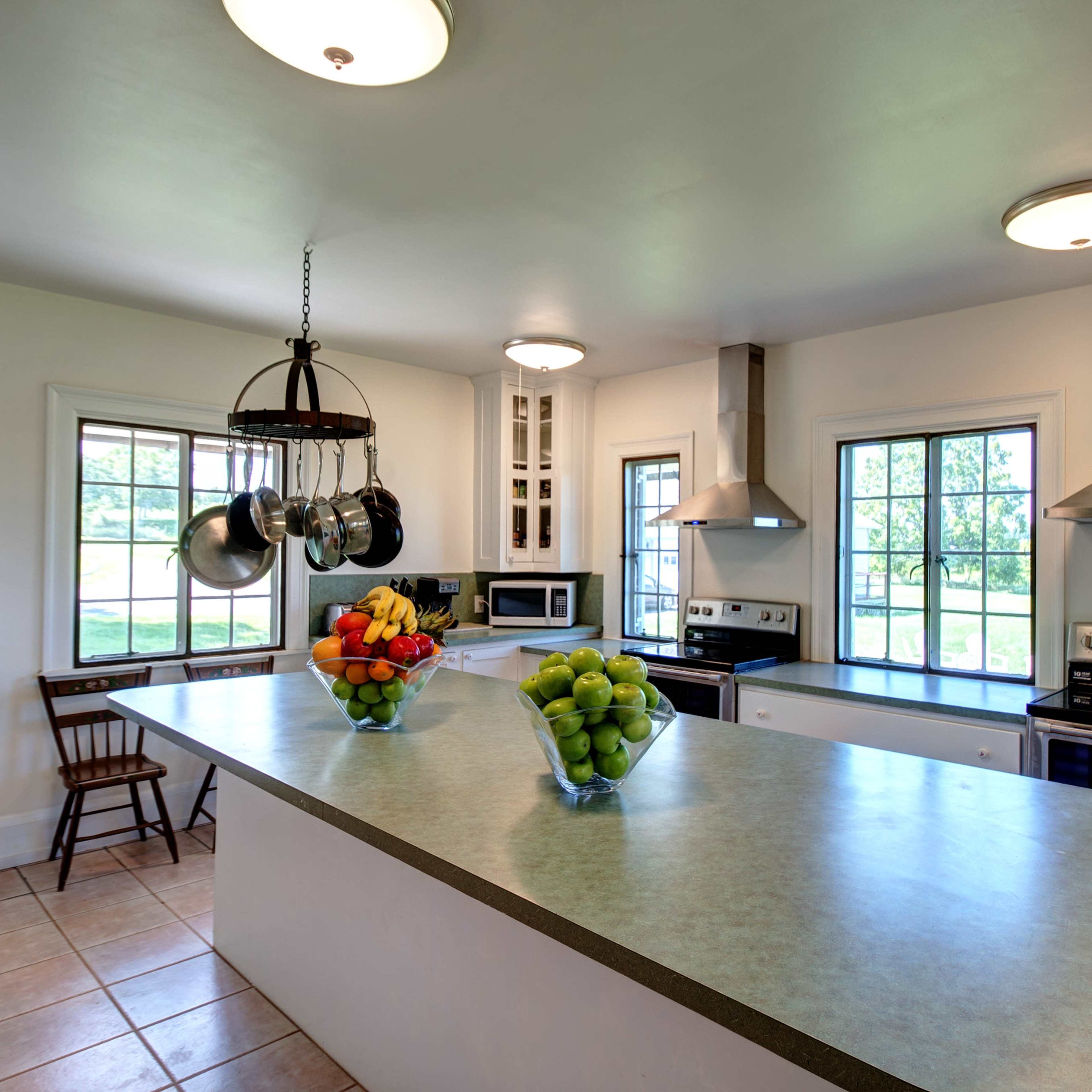 The kitchen features a central island, stainless steel appliances, a bowl of fruit, and large windows that provide natural light.