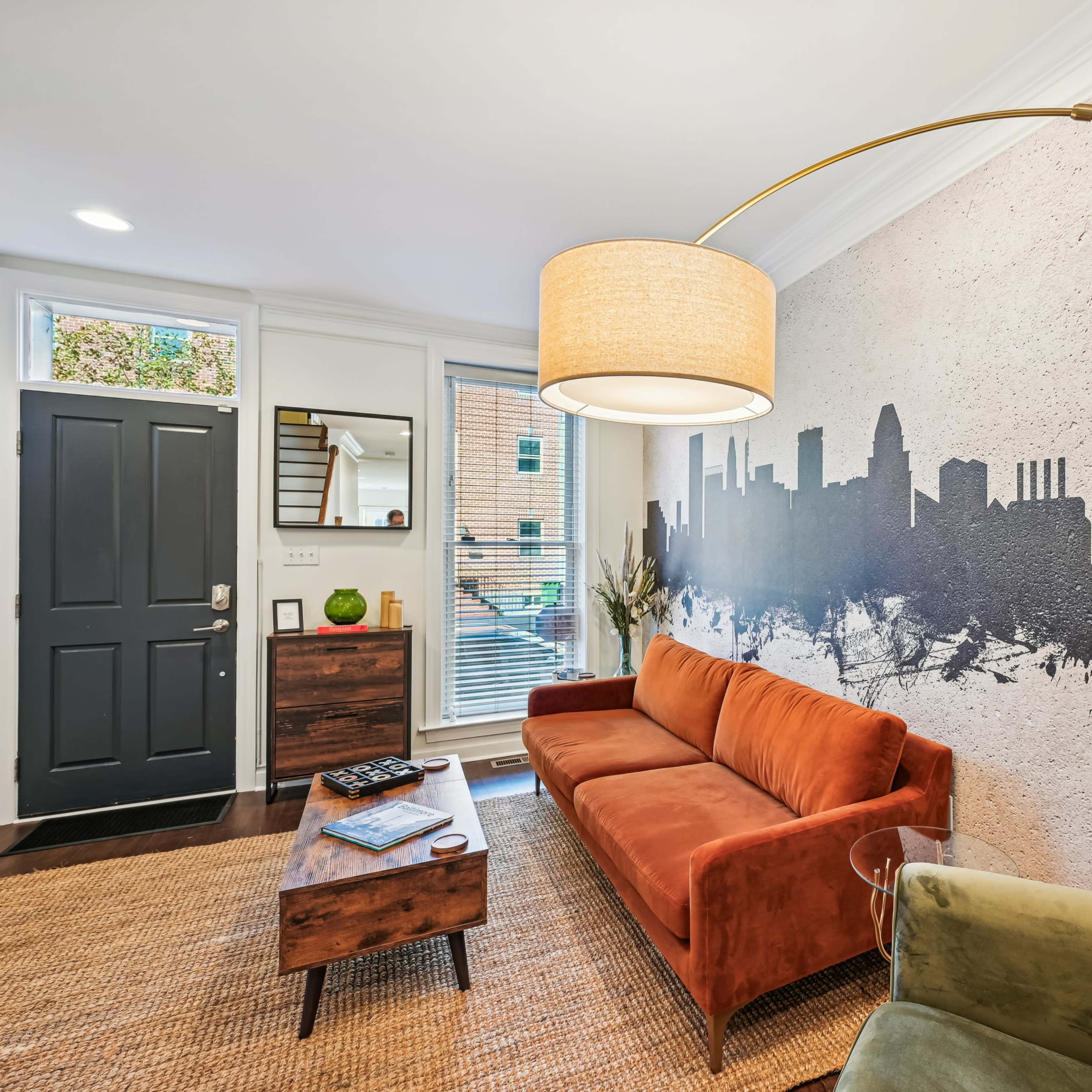 A living room features an orange sofa, a green chair, and a large wall mural of a city skyline.