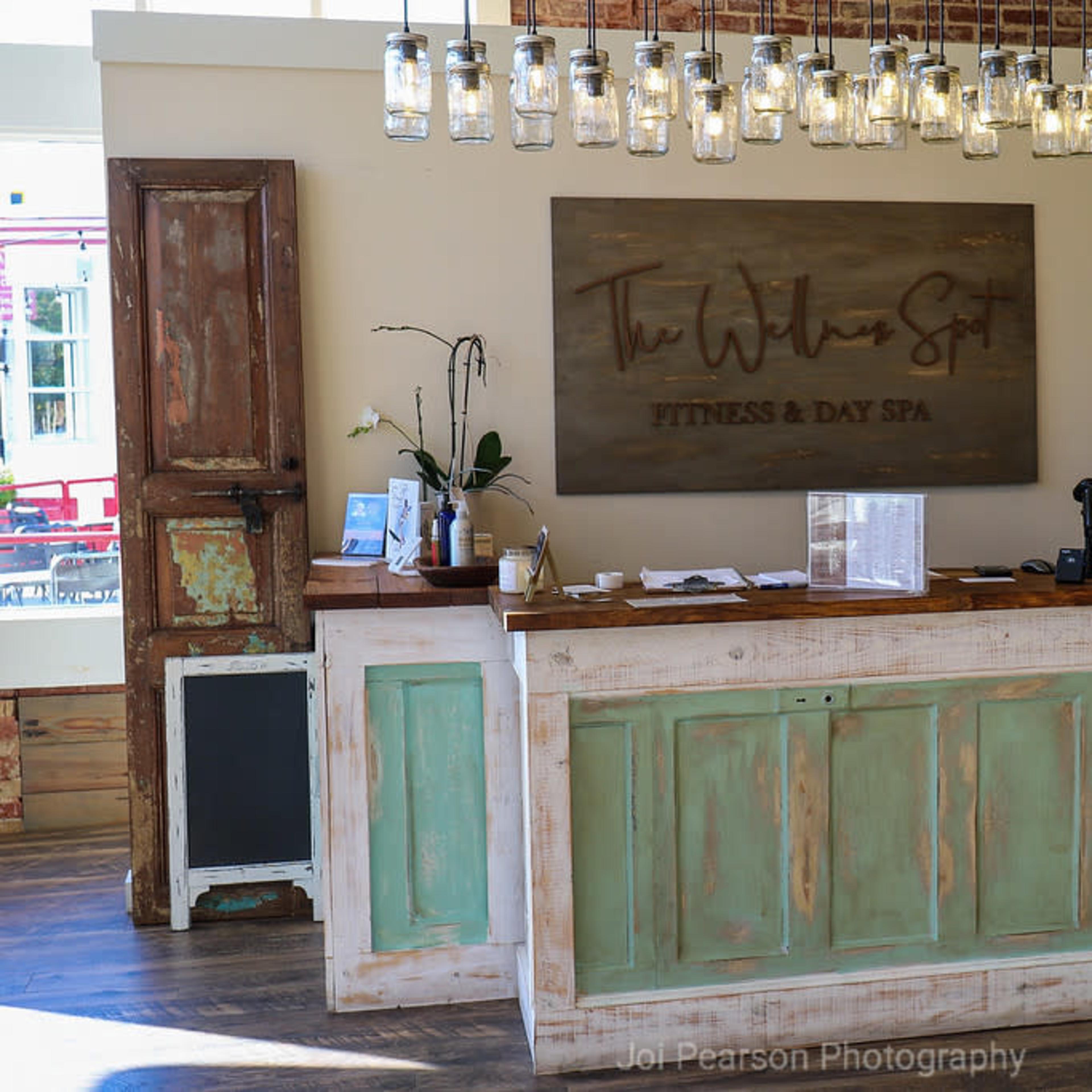 A reception desk at a spa, featuring a rustic design with green cabinetry, a large wooden sign, and hanging light fixtures.