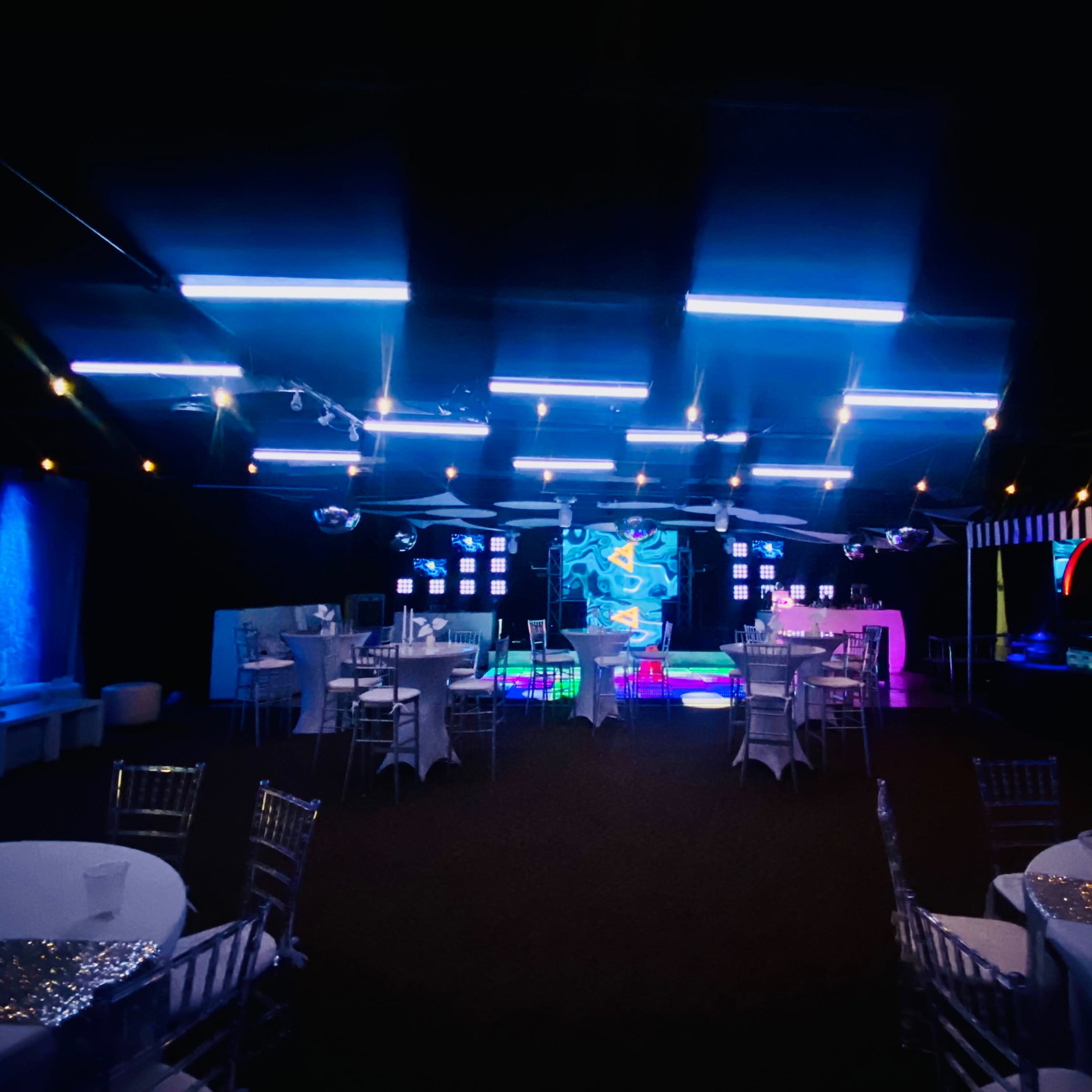 A dimly lit event space with tables and chairs arranged around a colorful backdrop featuring neon lights and screens.