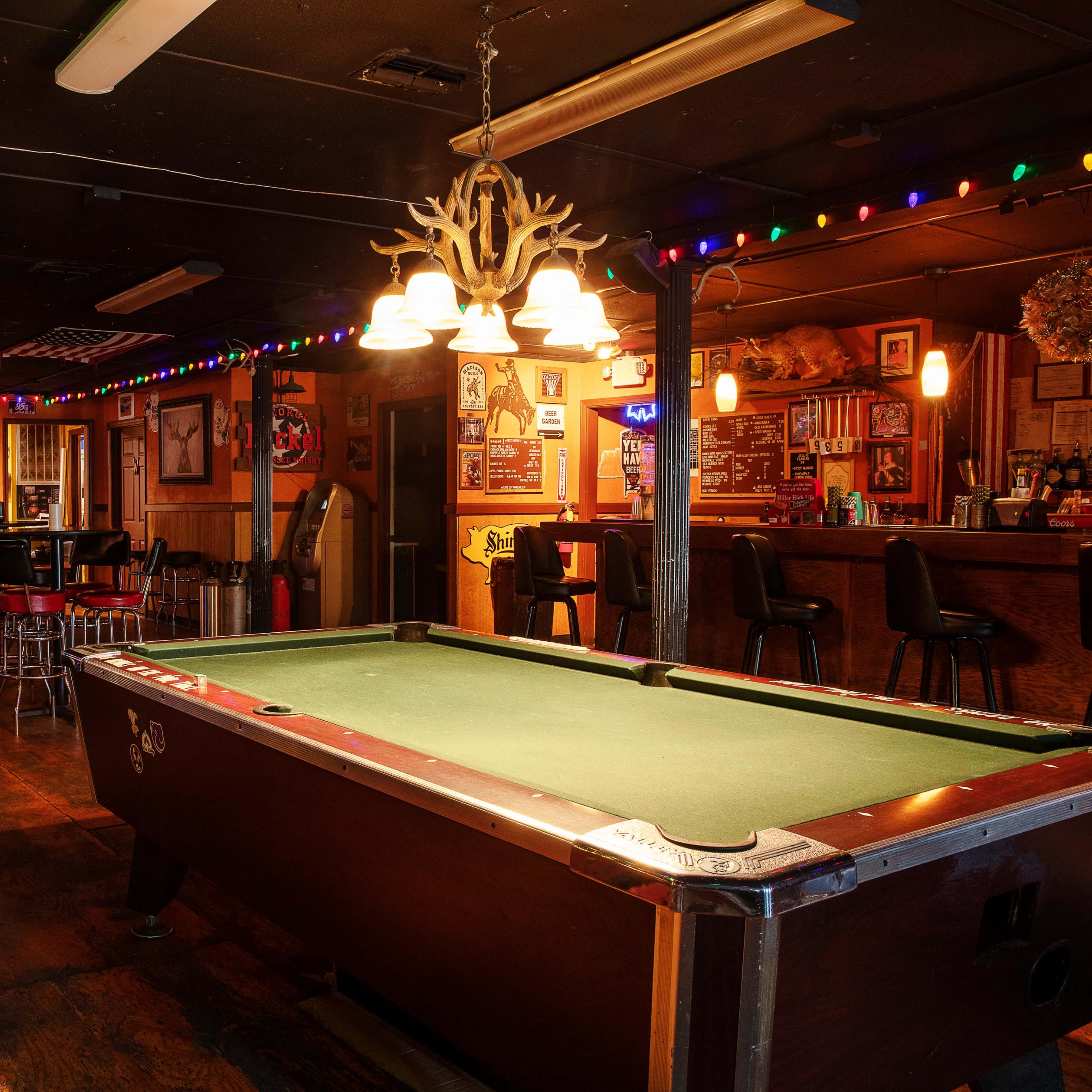 A dimly lit bar featuring a pool table, colorful string lights, and a wooden counter with stools.