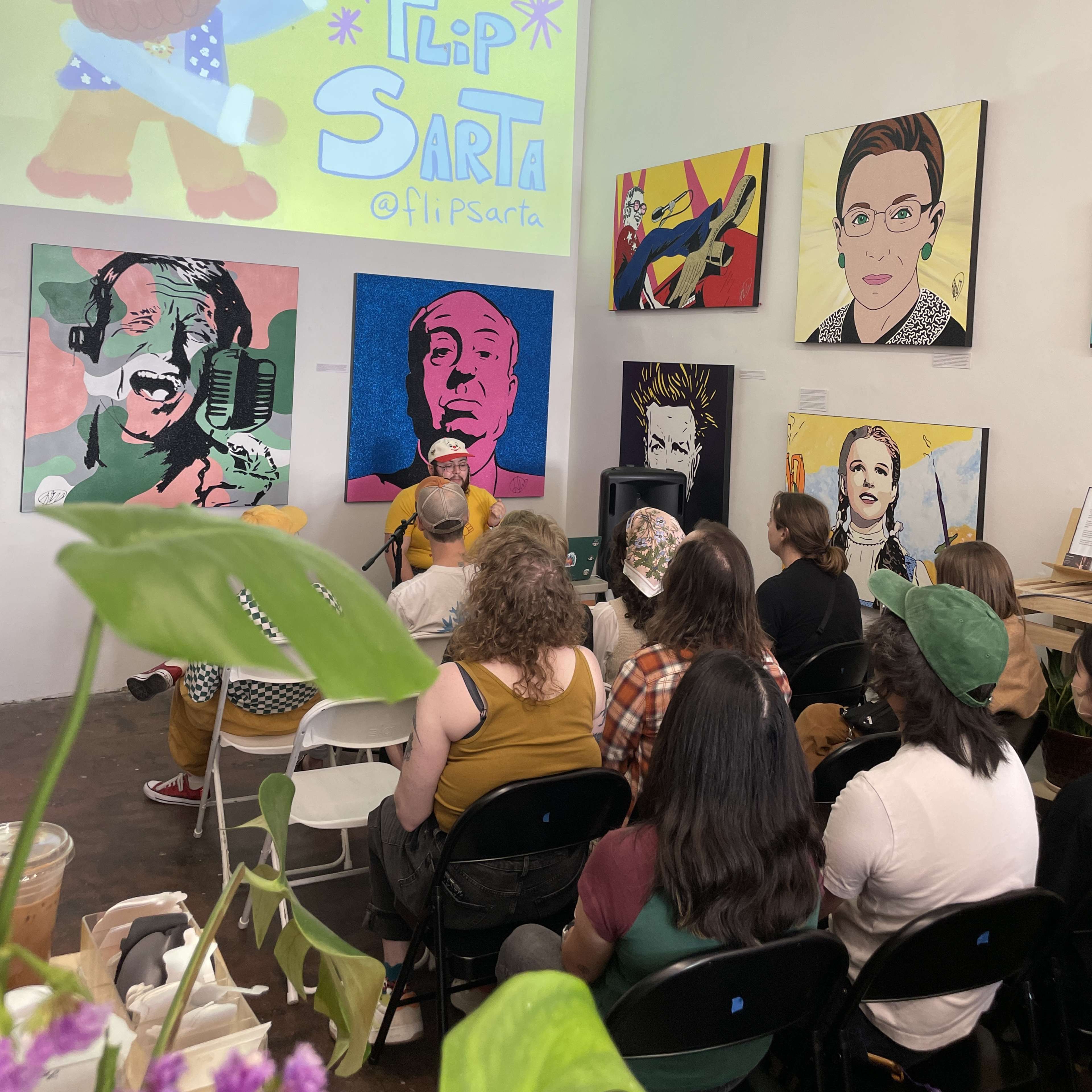 A group of people sits in front of colorful artwork on the walls, while a presentation titled "Flip Sarta" is projected on a screen.