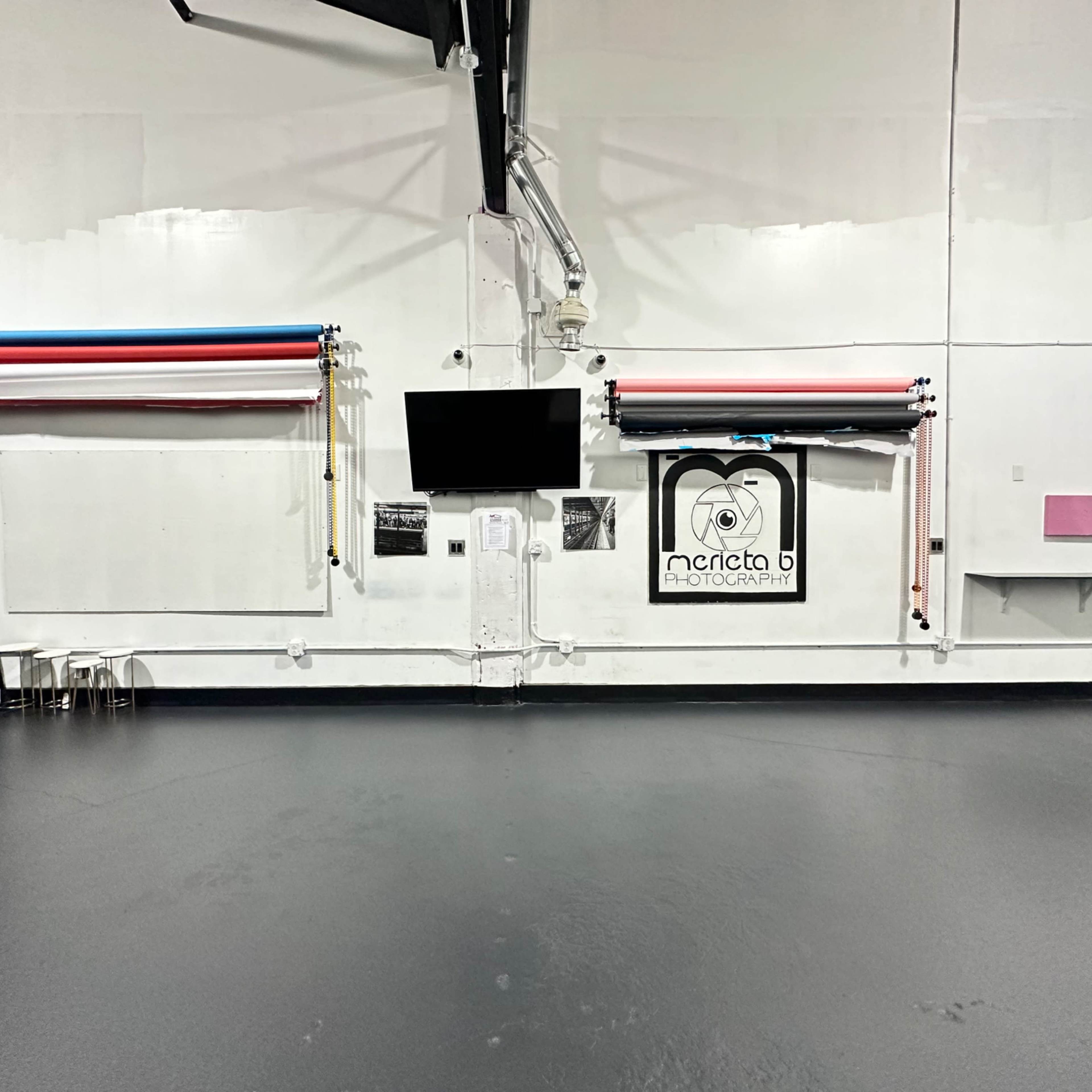 A photography studio with a blank wall, a TV mounted on it, backdrop rolls in various colors, and some chairs positioned on the left.
