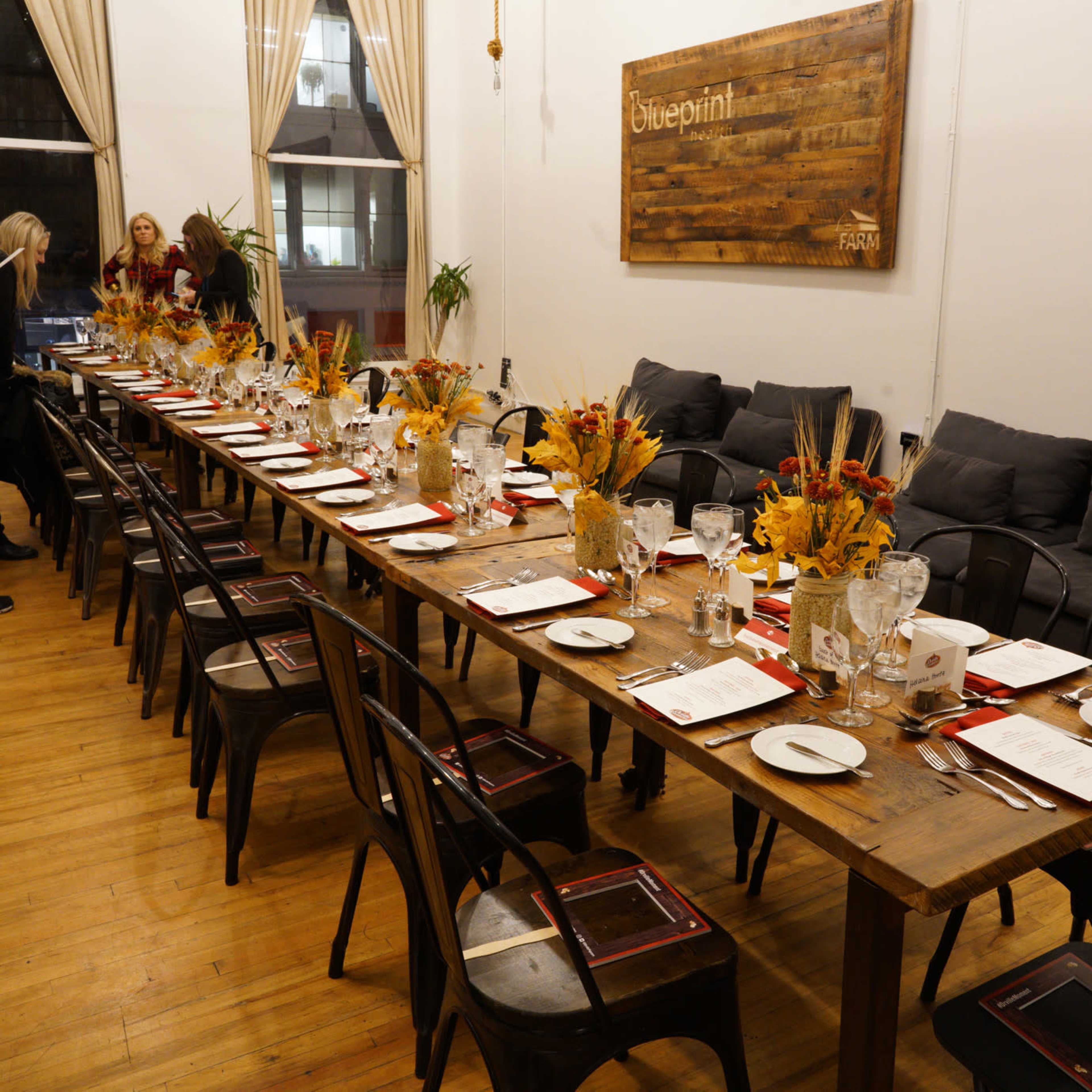 A long wooden dining table is set with plates and glasses, adorned with floral centerpieces, in a room featuring large windows and comfortable seating.