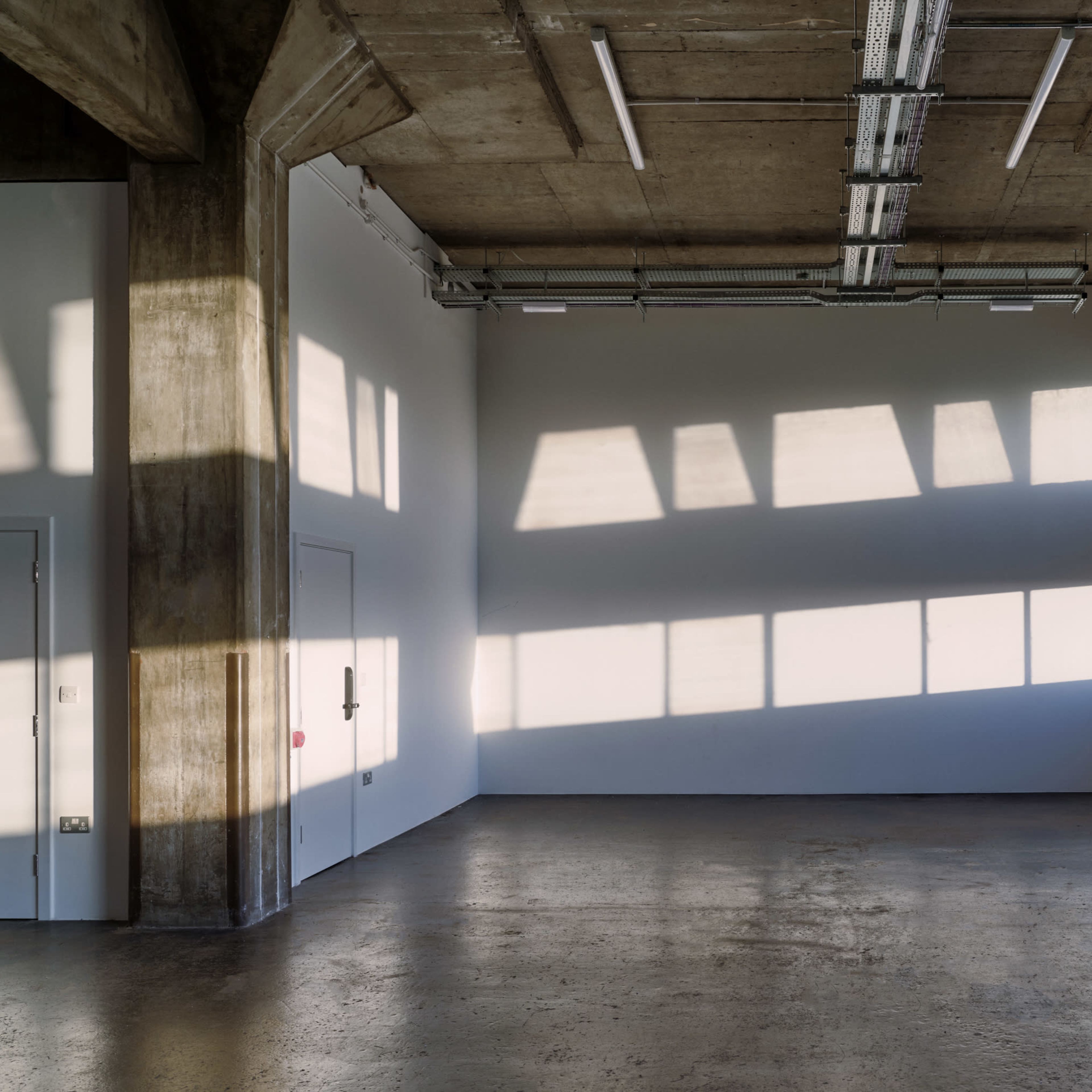 A spacious room with concrete walls and large windows casting shadows on the floor.