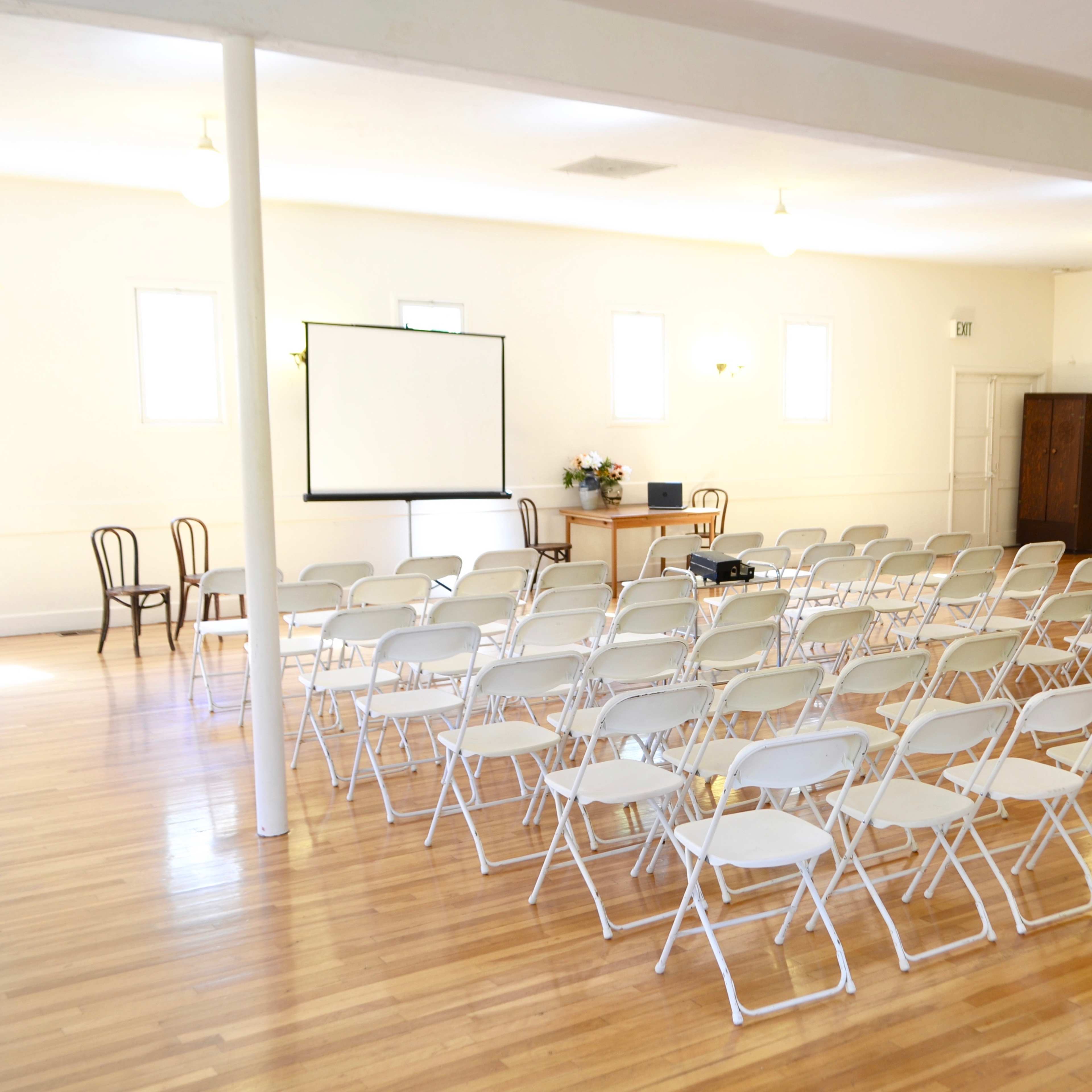 A spacious, empty room set up with rows of white folding chairs facing a projector screen, with a table and a flower arrangement in the background.
