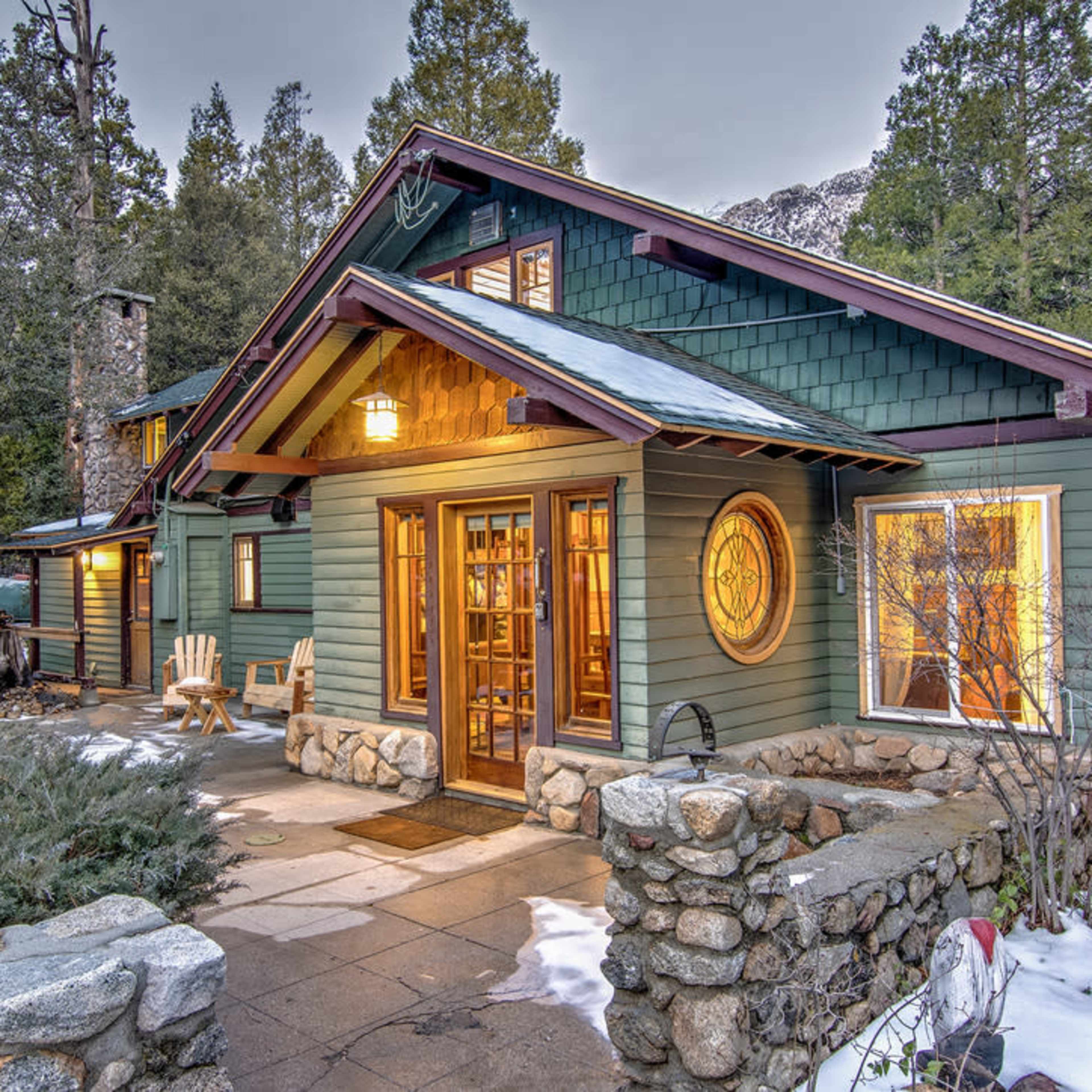 A stone and wood cabin with large windows and a welcoming porch is surrounded by evergreen trees and patches of snow.