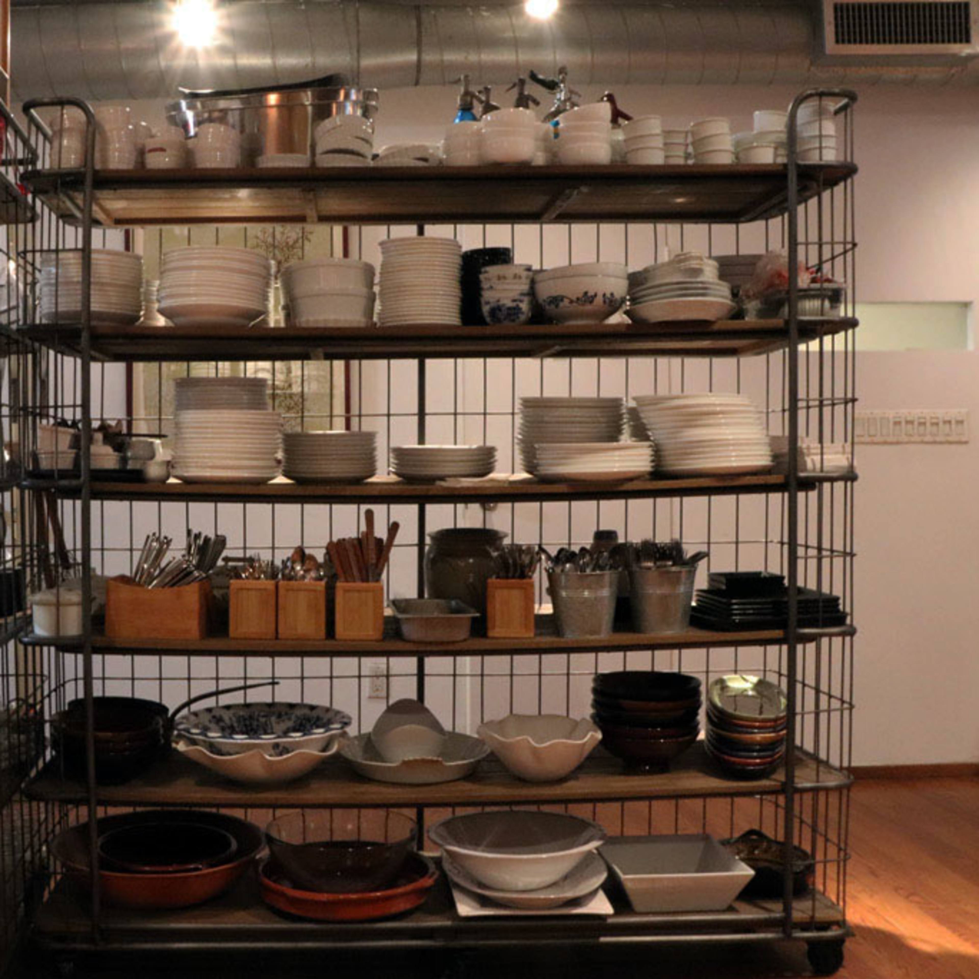 A metal shelving unit displays an assortment of plates, bowls, utensils, and kitchen tools in a kitchen setting.