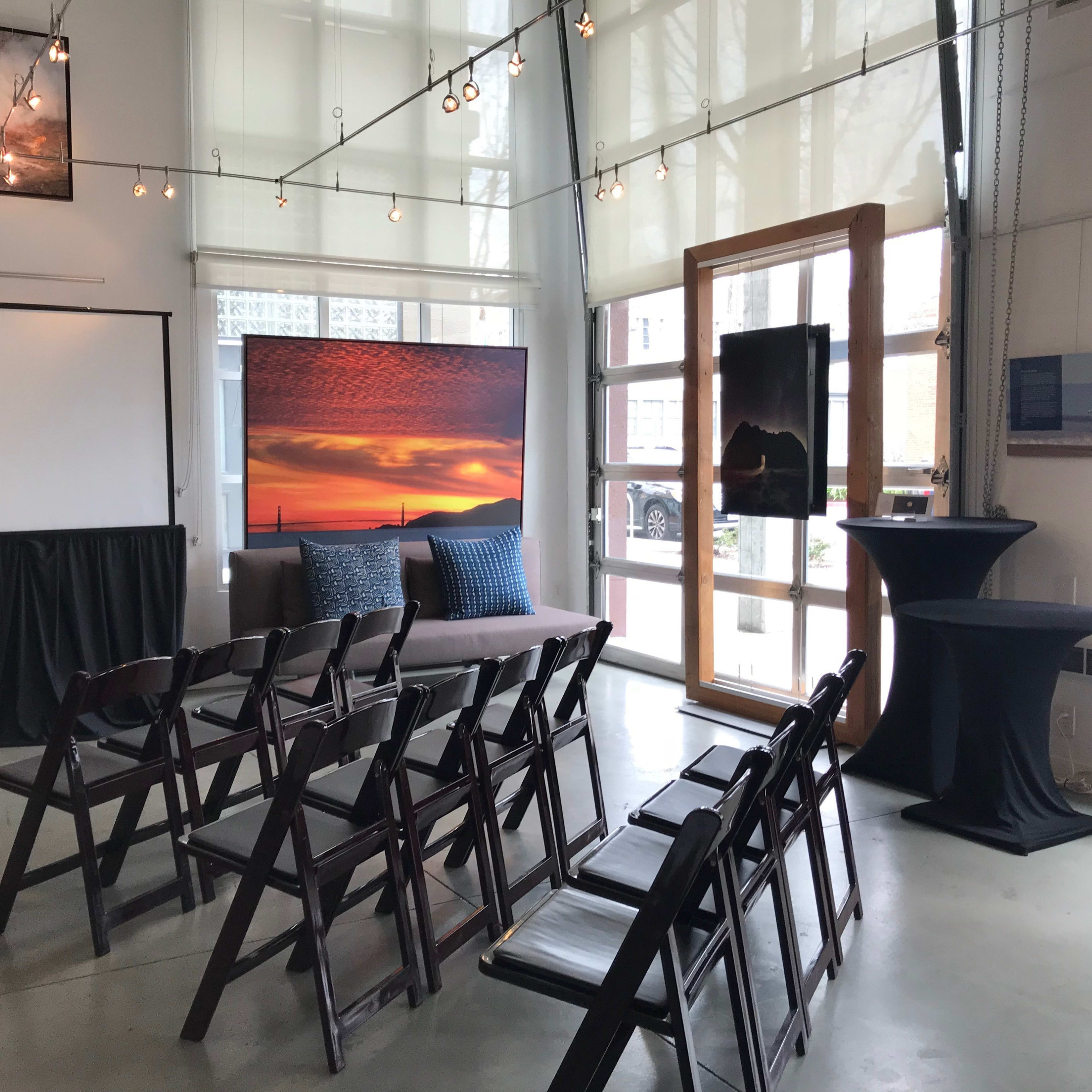 A modern event space set up with rows of folding chairs facing a blank screen, framed by large windows and decorated with artwork on the walls.