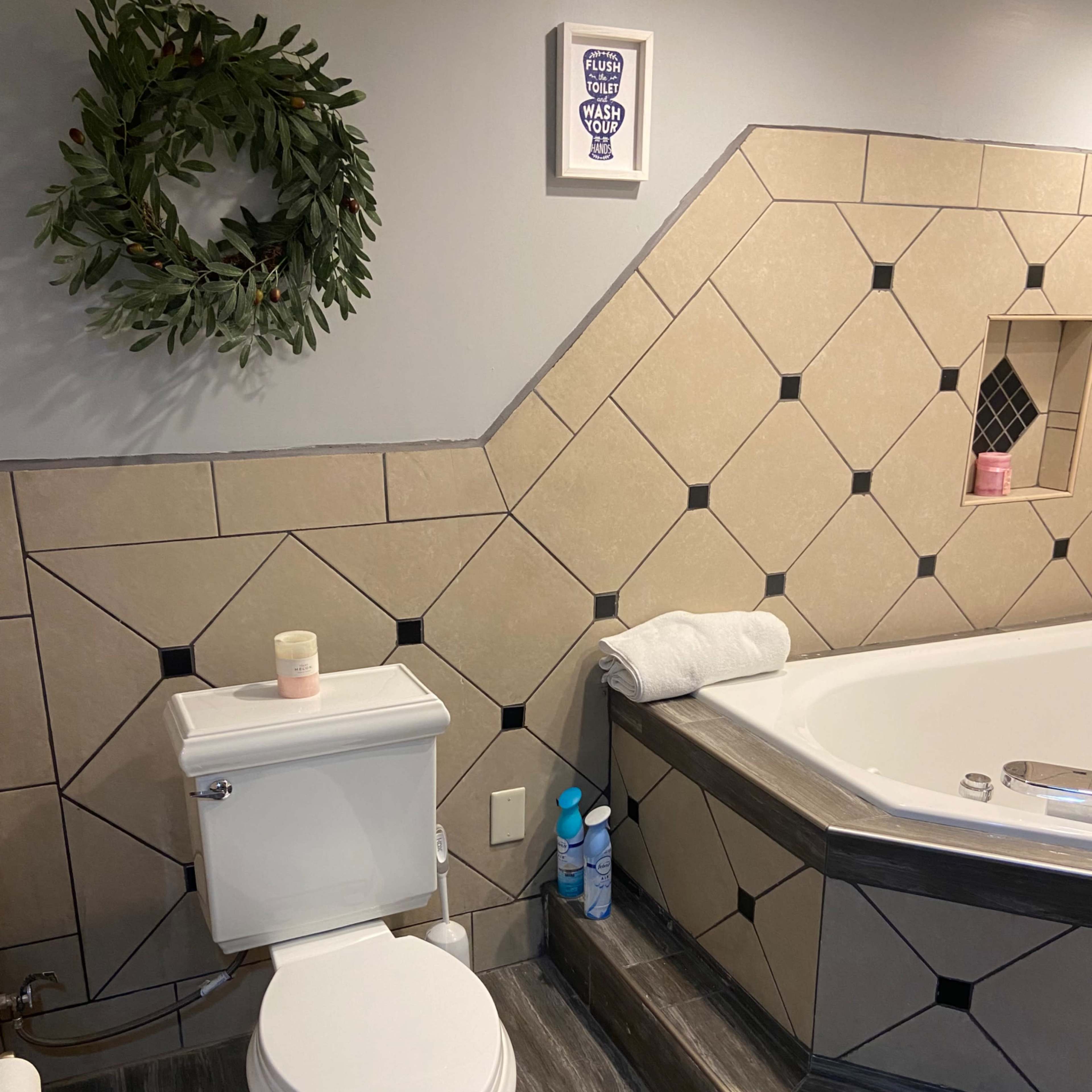 A bathroom featuring a toilet next to a large bathtub with decorative tiles and a small wreath on the wall.