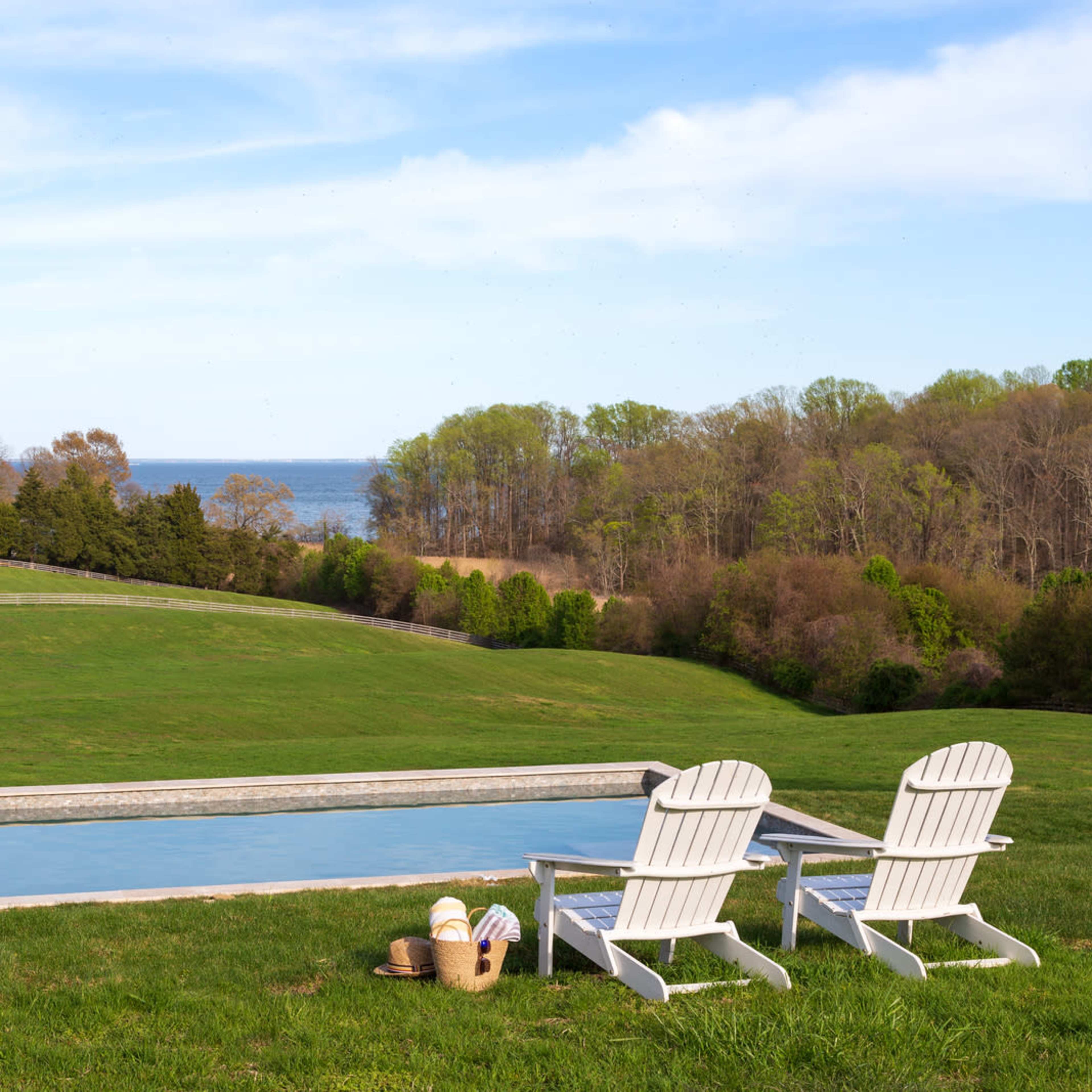Two white Adirondack chairs sit beside a long, narrow swimming pool overlooking a grassy field and a body of water in the distance.