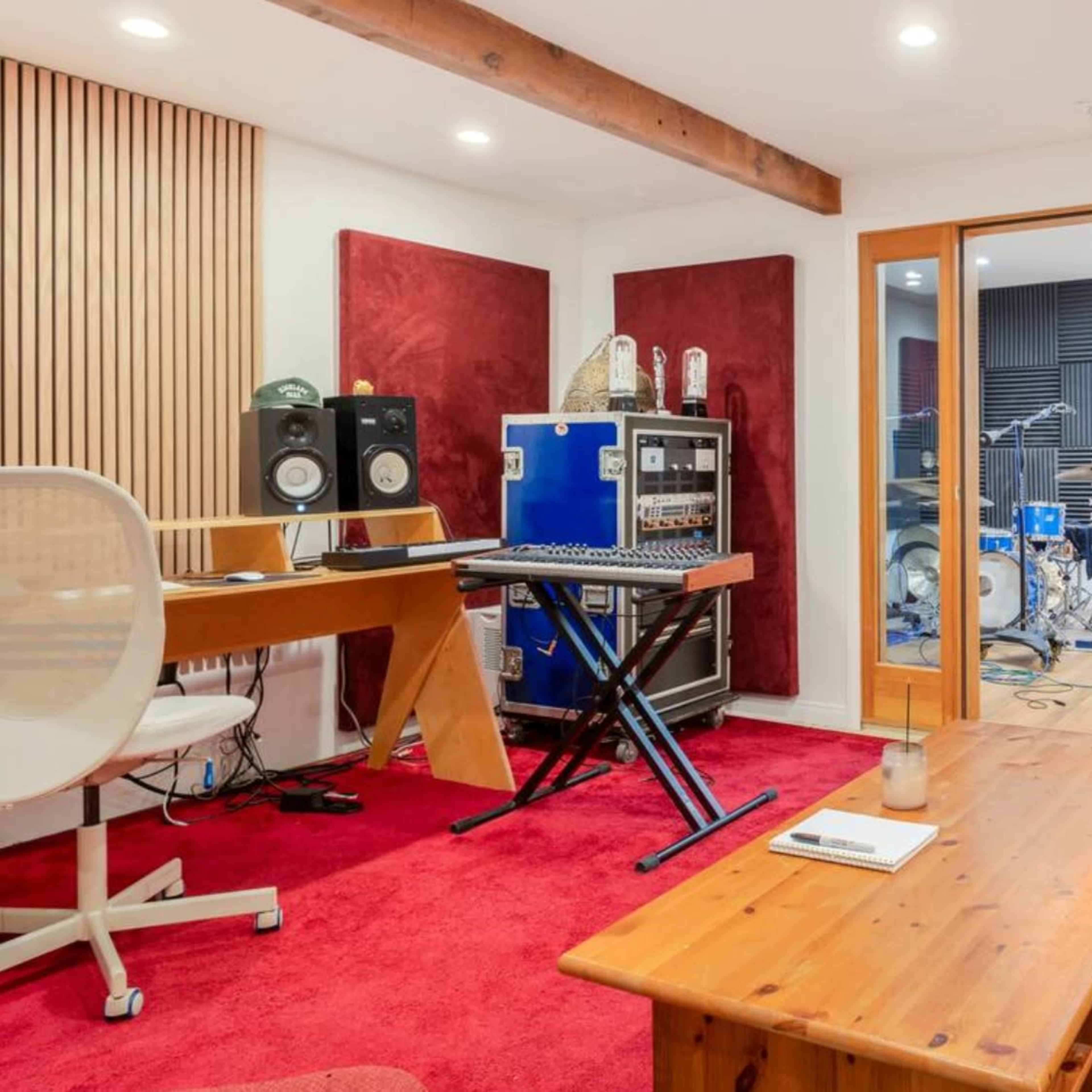 A music recording studio with a keyboard, audio equipment, and a visible drum set through glass doors.