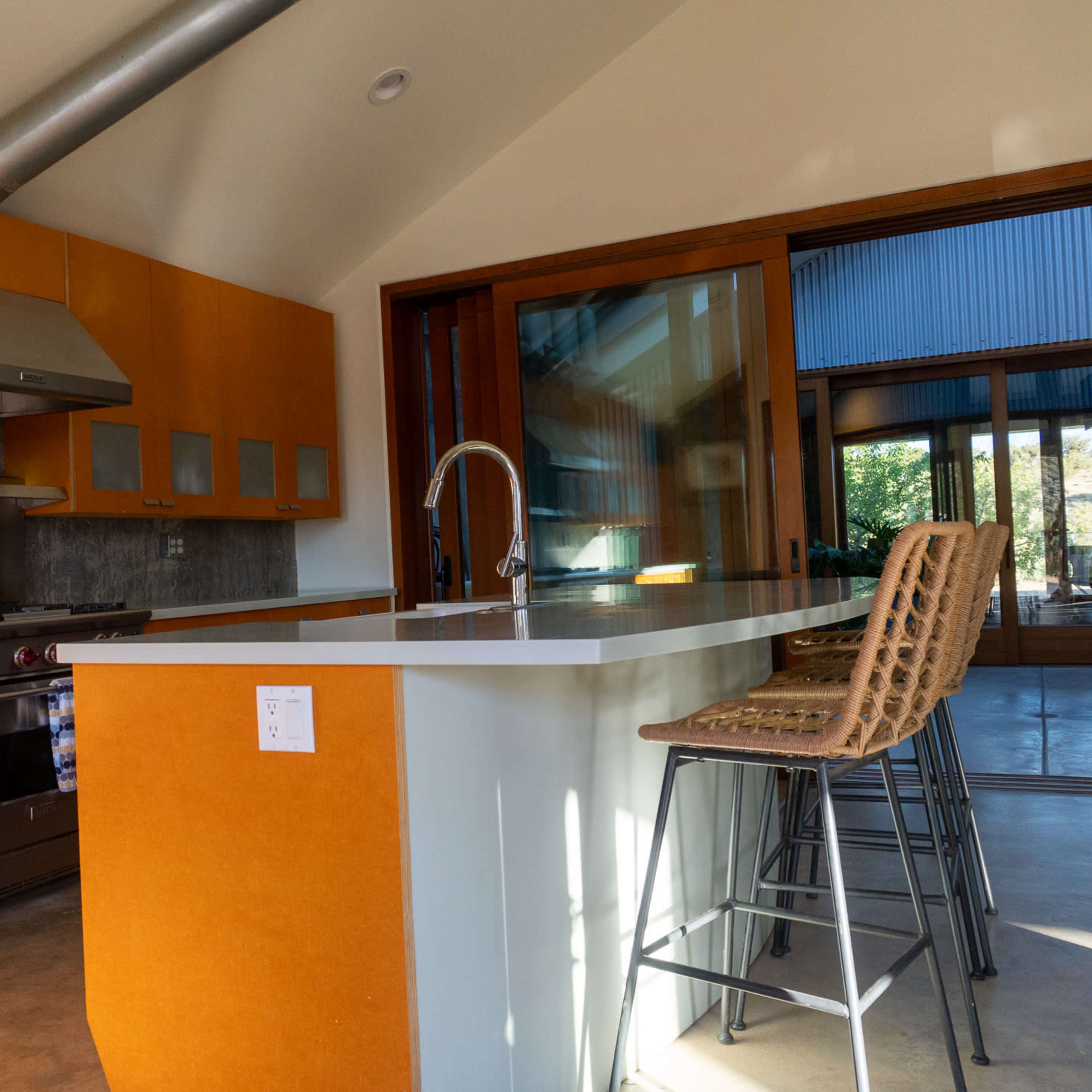 A modern kitchen with orange cabinetry, stainless steel appliances, and bar stools at a white countertop.