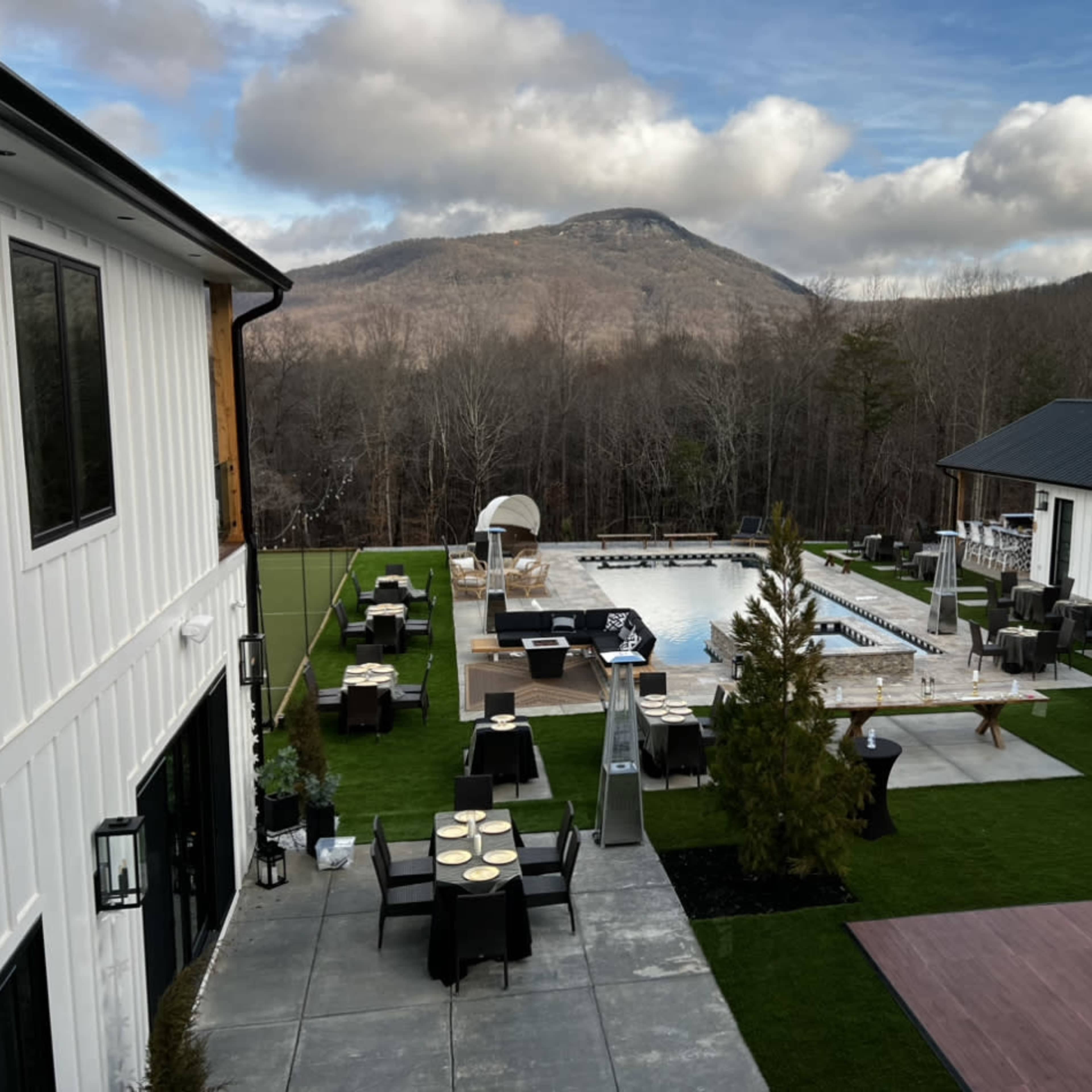 A modern outdoor area featuring a large pool, lounge furniture, and tables, surrounded by greenery and mountains in the background.