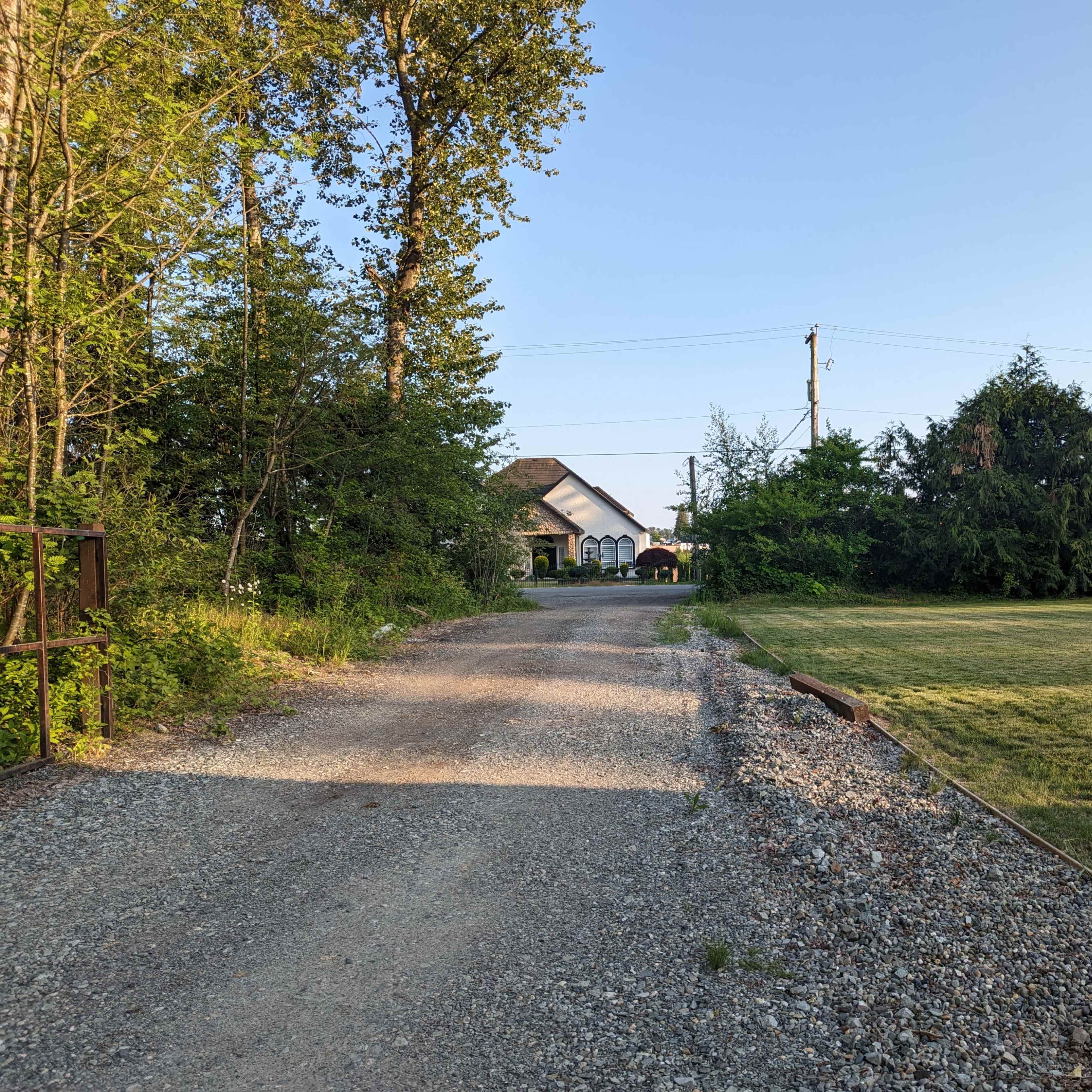 A gravel pathway leads toward a house surrounded by trees and a grassy area under a clear sky.