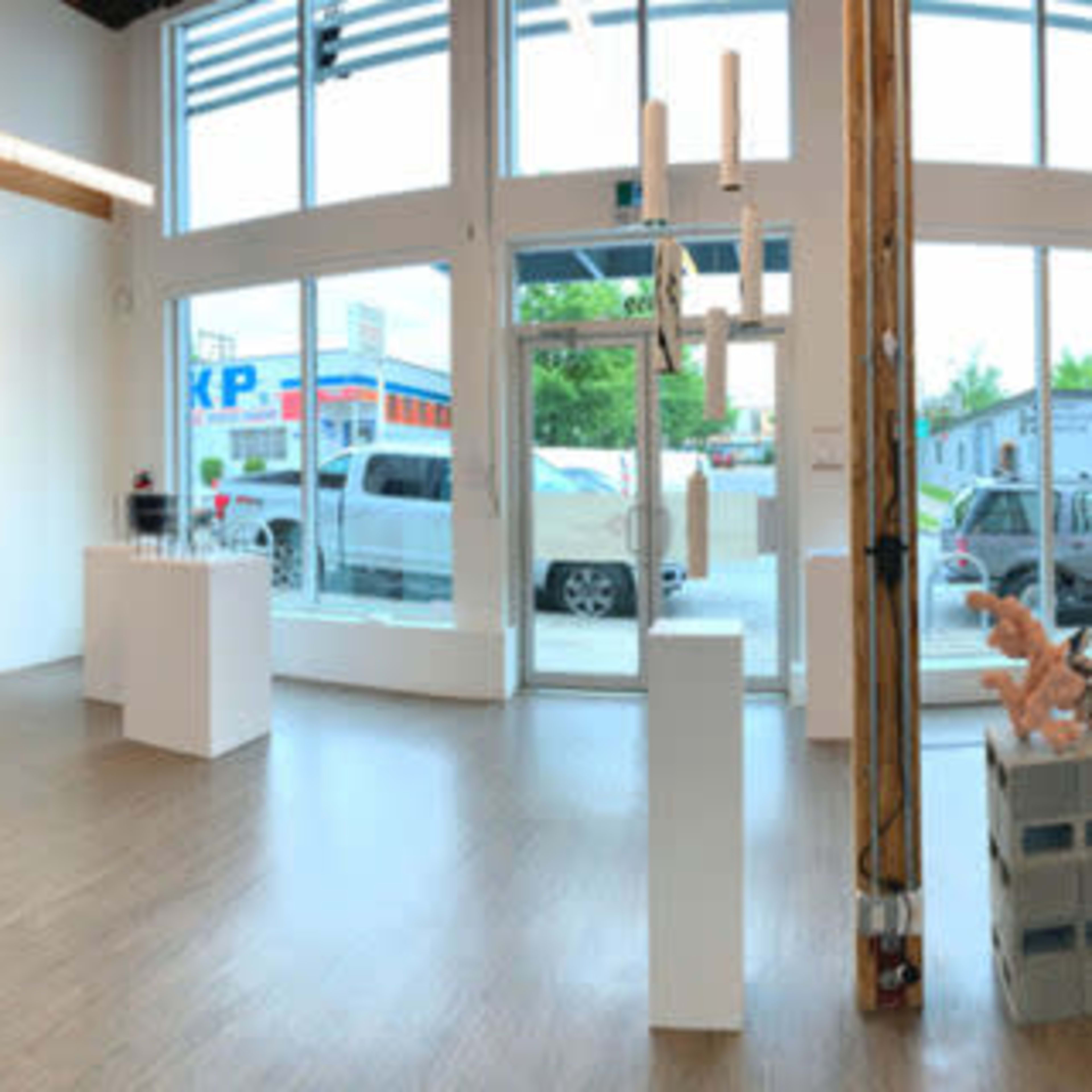 An art gallery interior featuring various sculptures and artworks displayed on white pedestals against a backdrop of large windows.