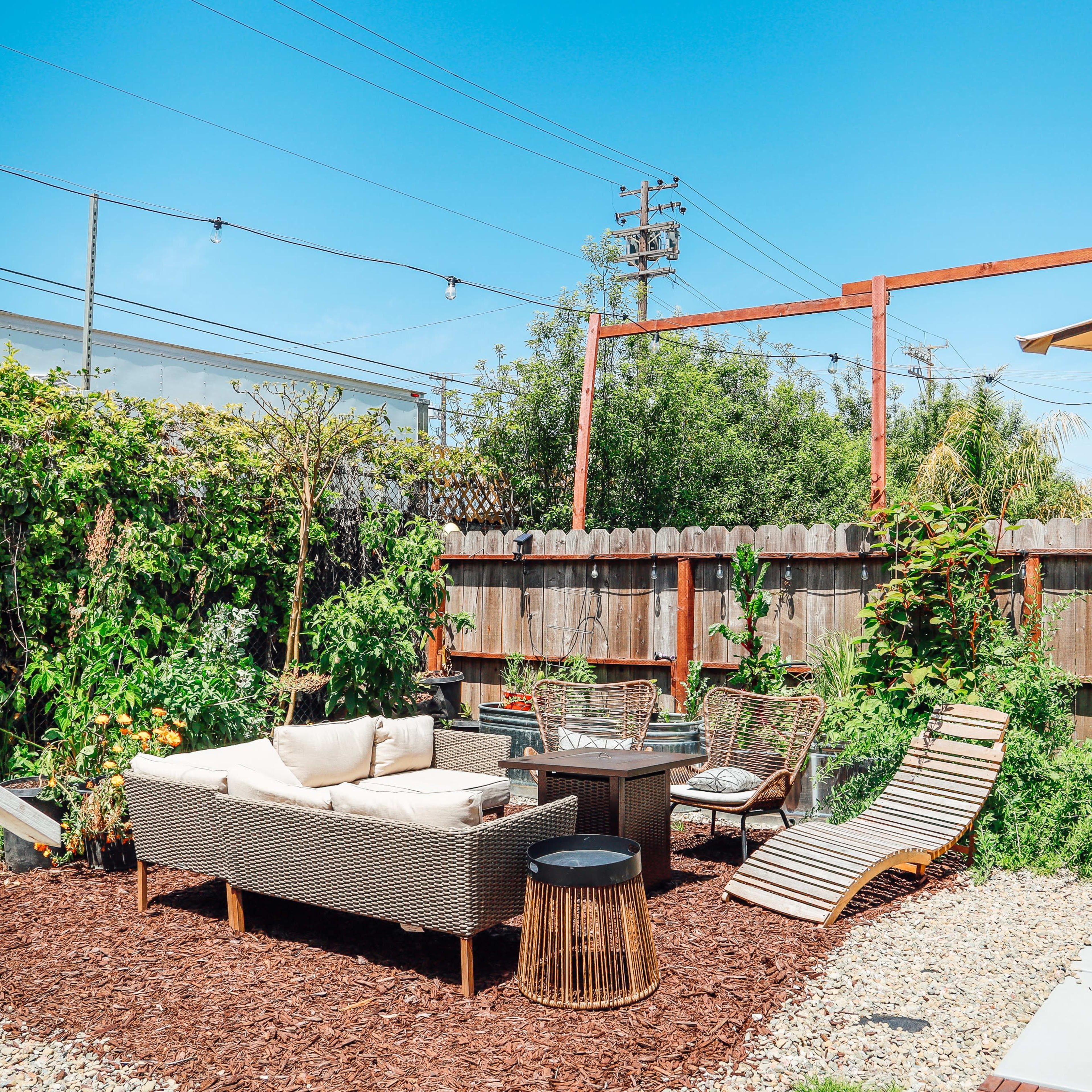 A landscaped outdoor space features a seating area with a sofa, chairs, and a lounge chair surrounded by greenery and a hot tub.