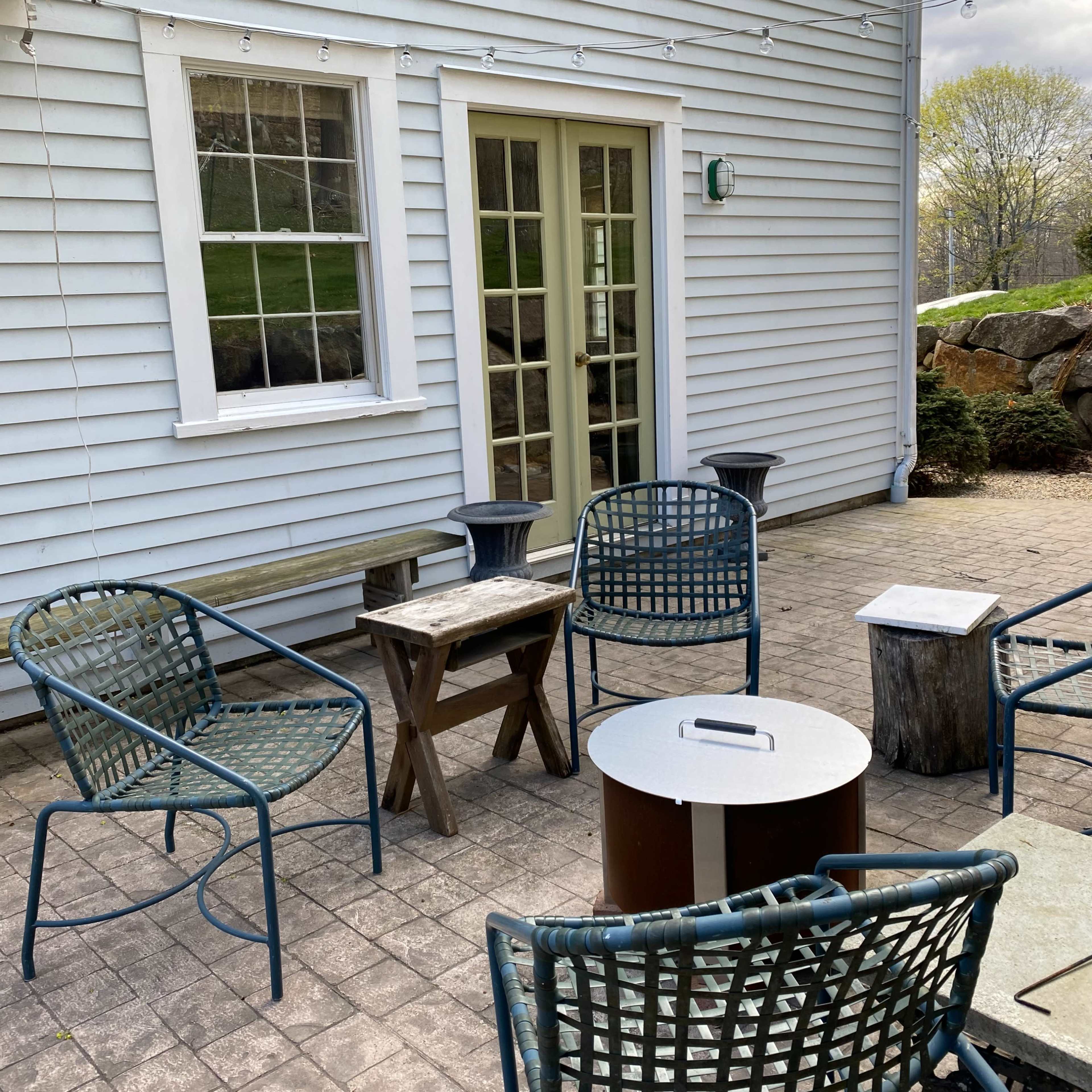 A patio area with several metal chairs, a wooden table, and a fire pit in front of a beige house with green doors and a stone wall nearby.
