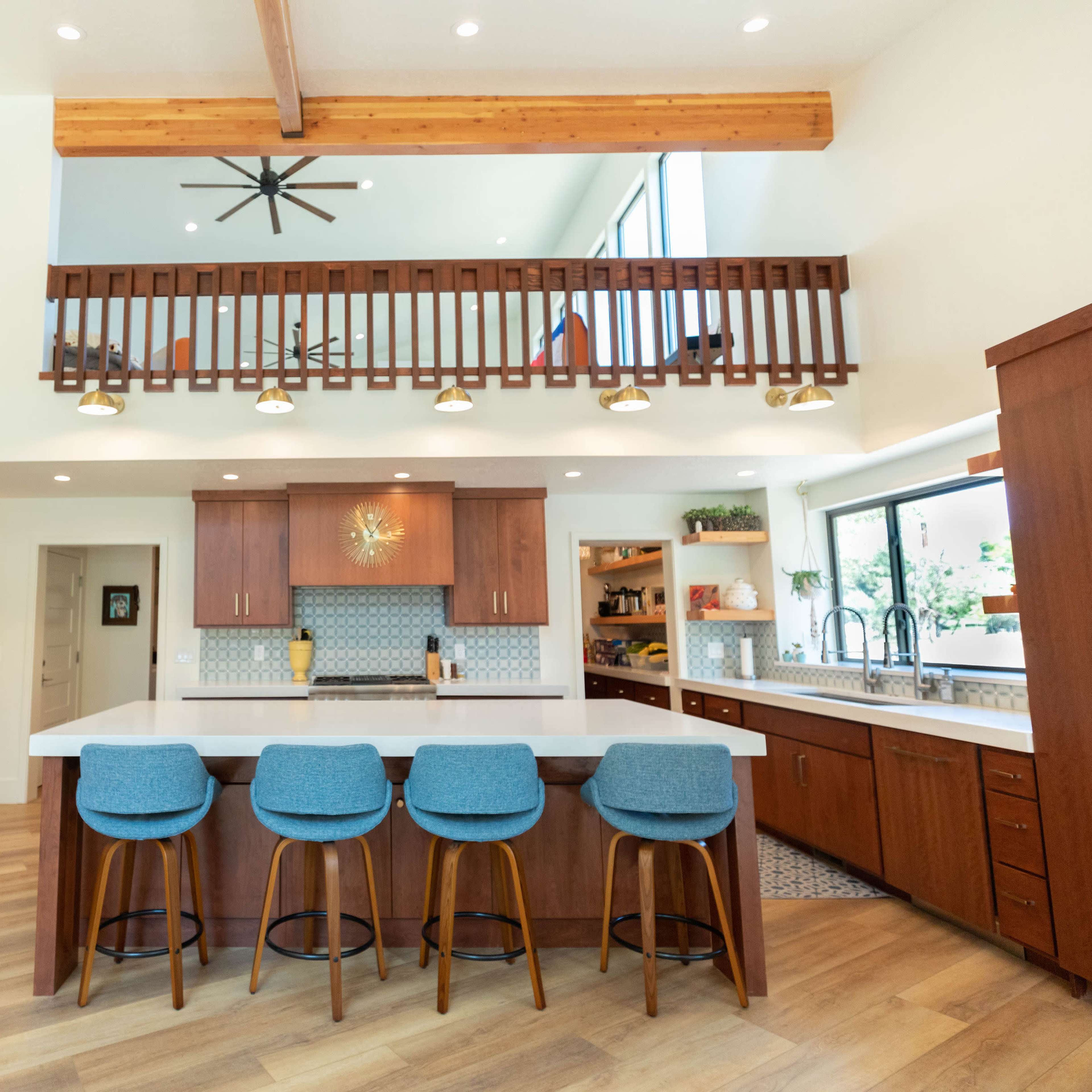 A modern kitchen with a central island and four blue bar stools, complemented by wooden cabinetry and an open design that leads to a balcony above.