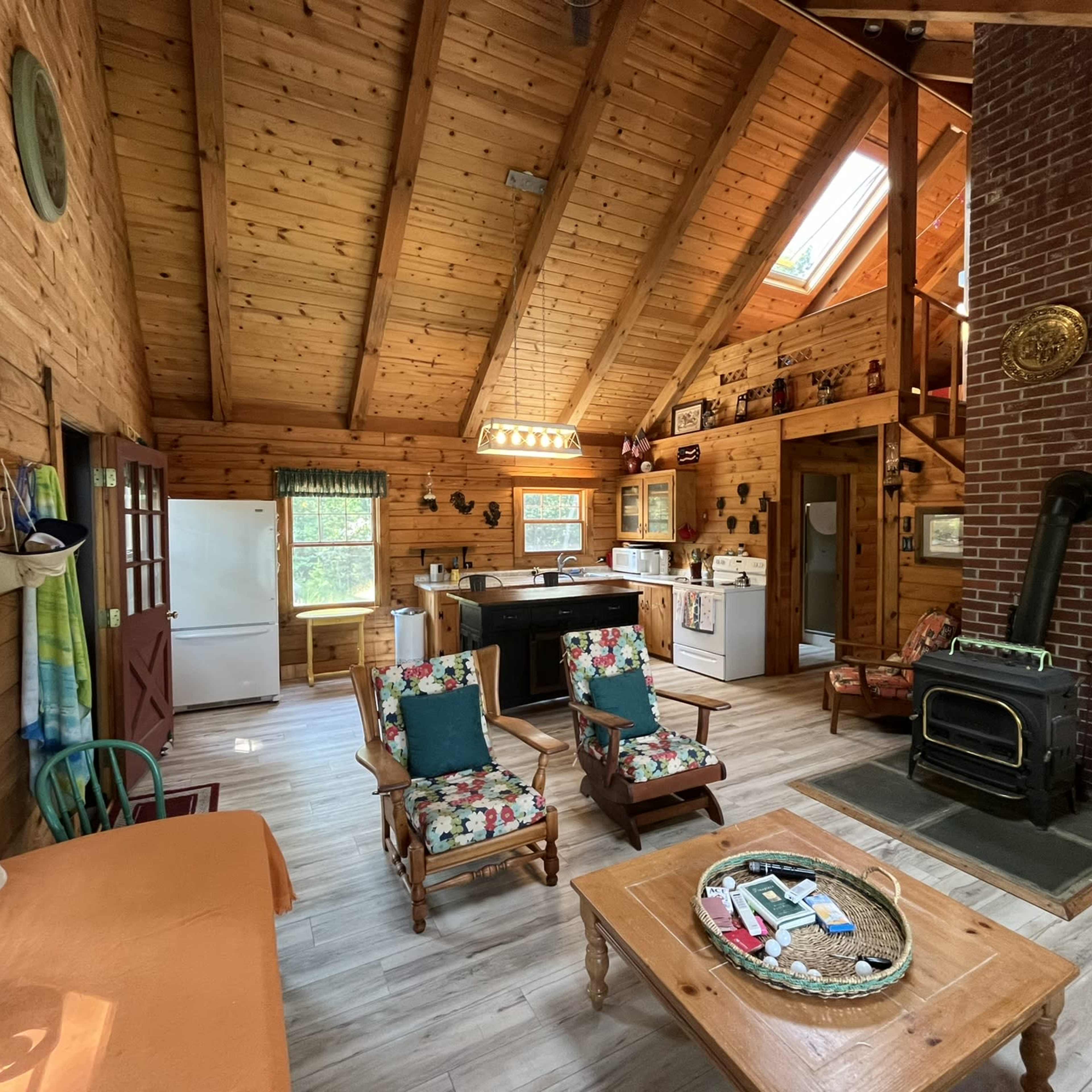 A rustic wooden cabin interior with a kitchen, living area, and a wood stove, featuring furniture arranged around a coffee table.