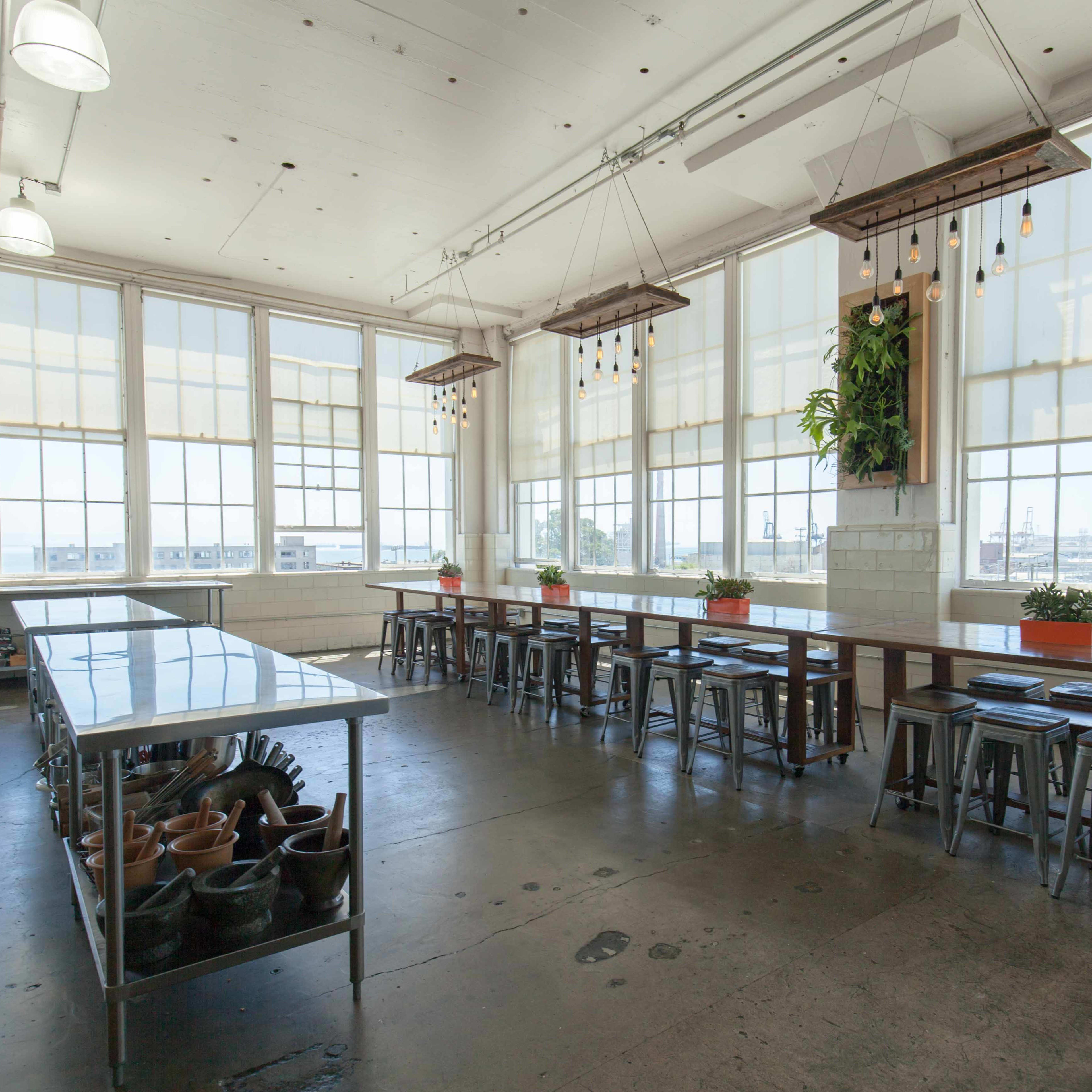 A spacious kitchen with large windows, long wooden tables, metal stools, and ceiling-mounted plants, all set against a concrete floor.