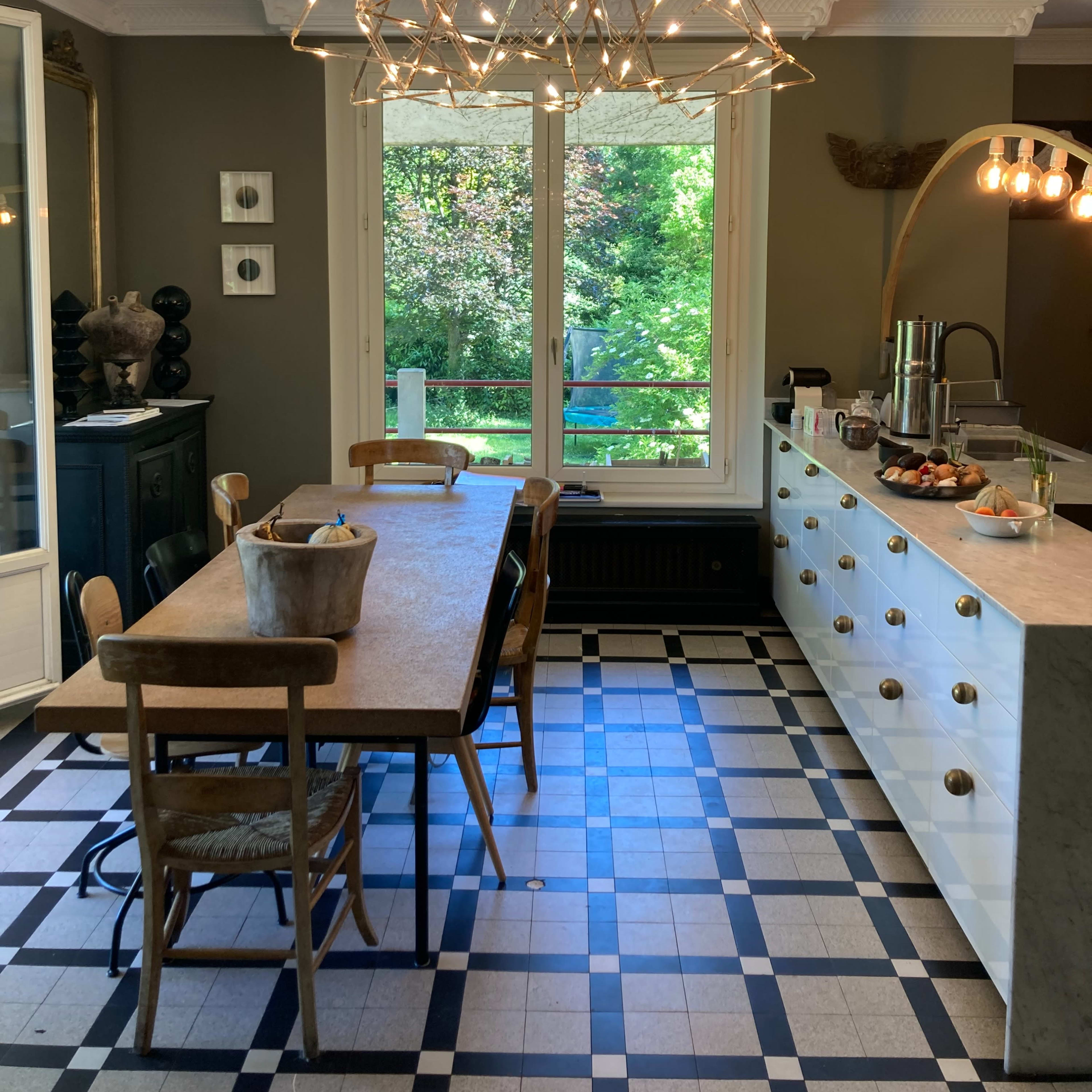 The kitchen features a large wooden dining table, a modern chandelier, and a view of a green garden through a set of windows.