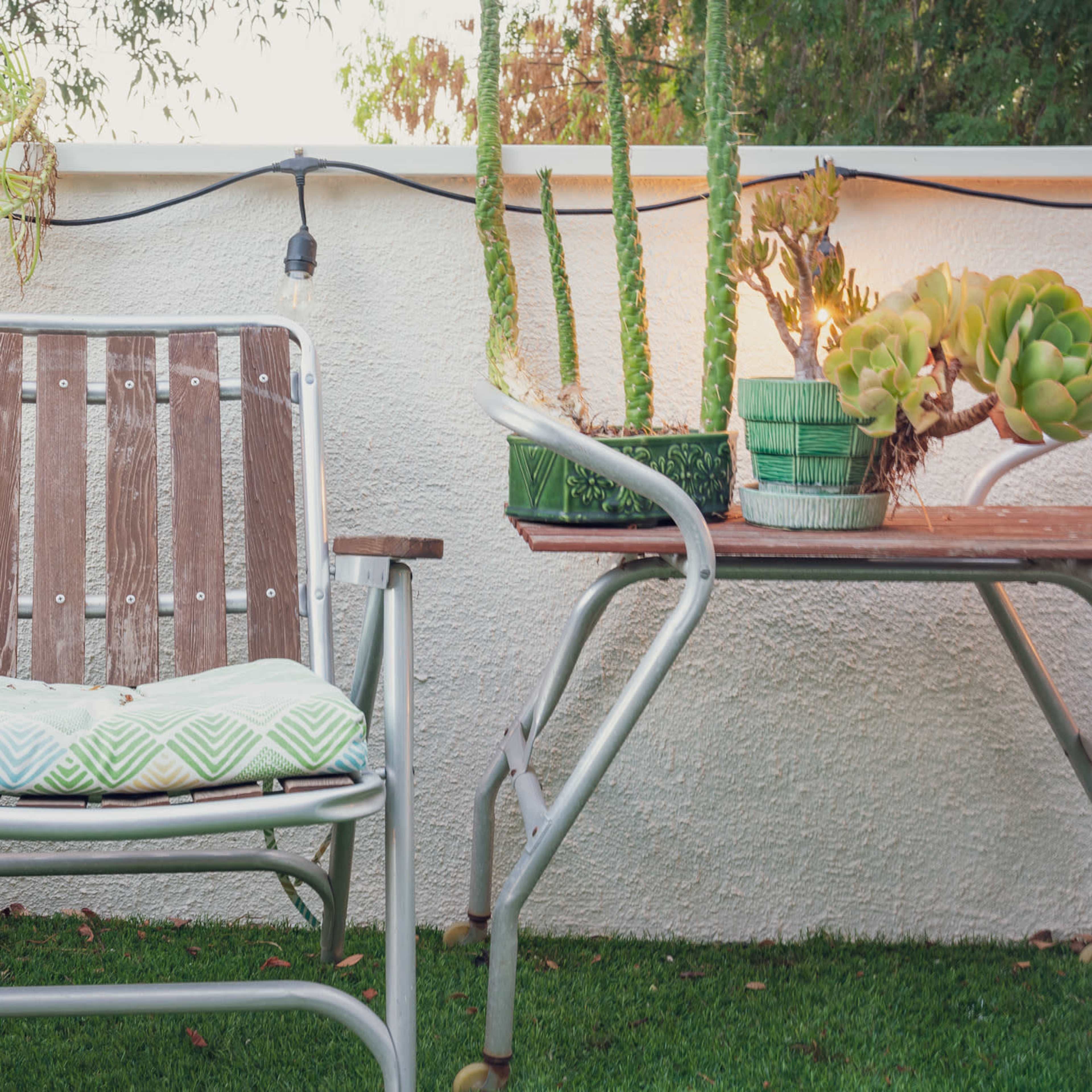 A metal outdoor chair with a patterned cushion sits beside a table adorned with potted succulents and string lights in a green garden space.