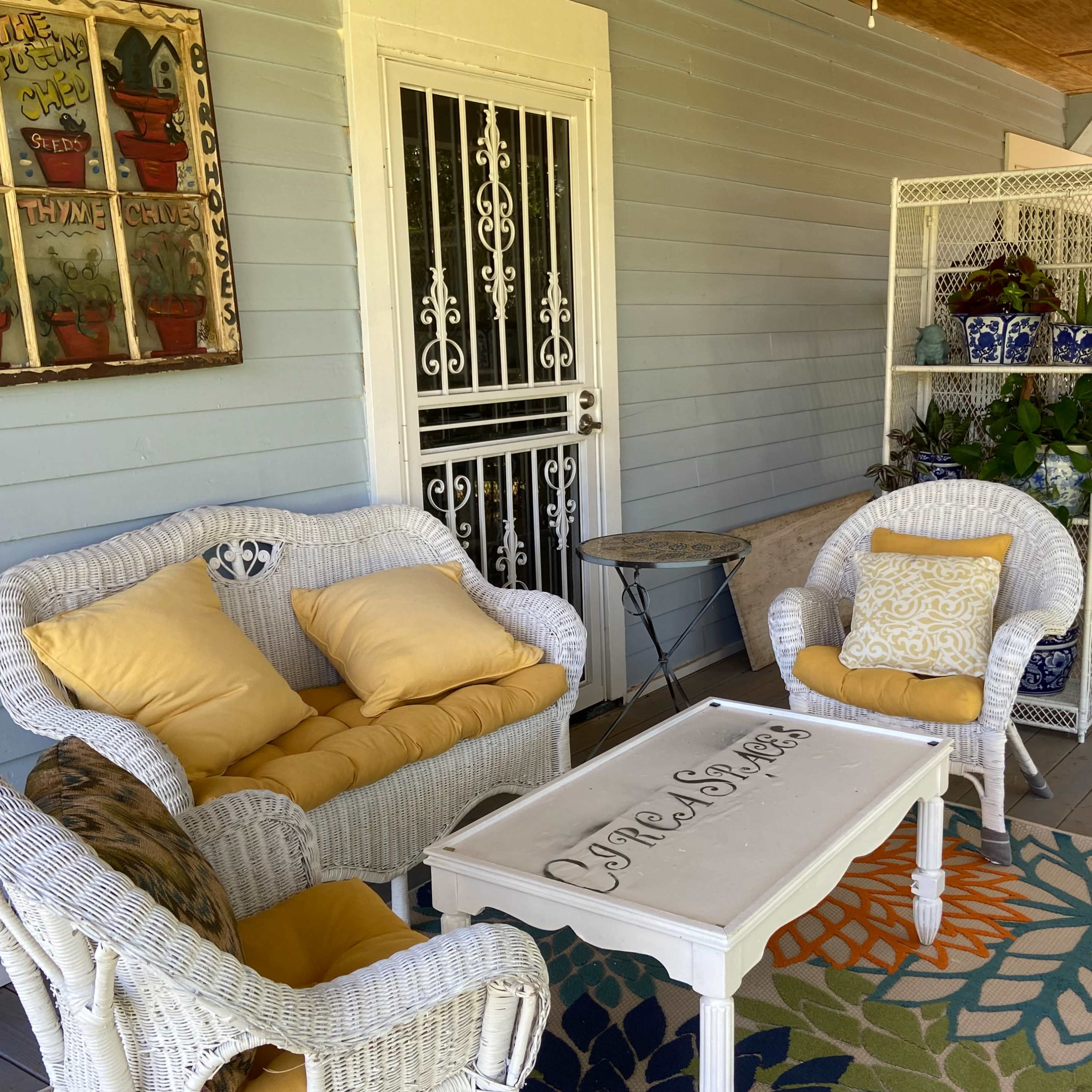 A cozy porch area with white wicker furniture, yellow cushions, a decorative table, and a variety of plants.