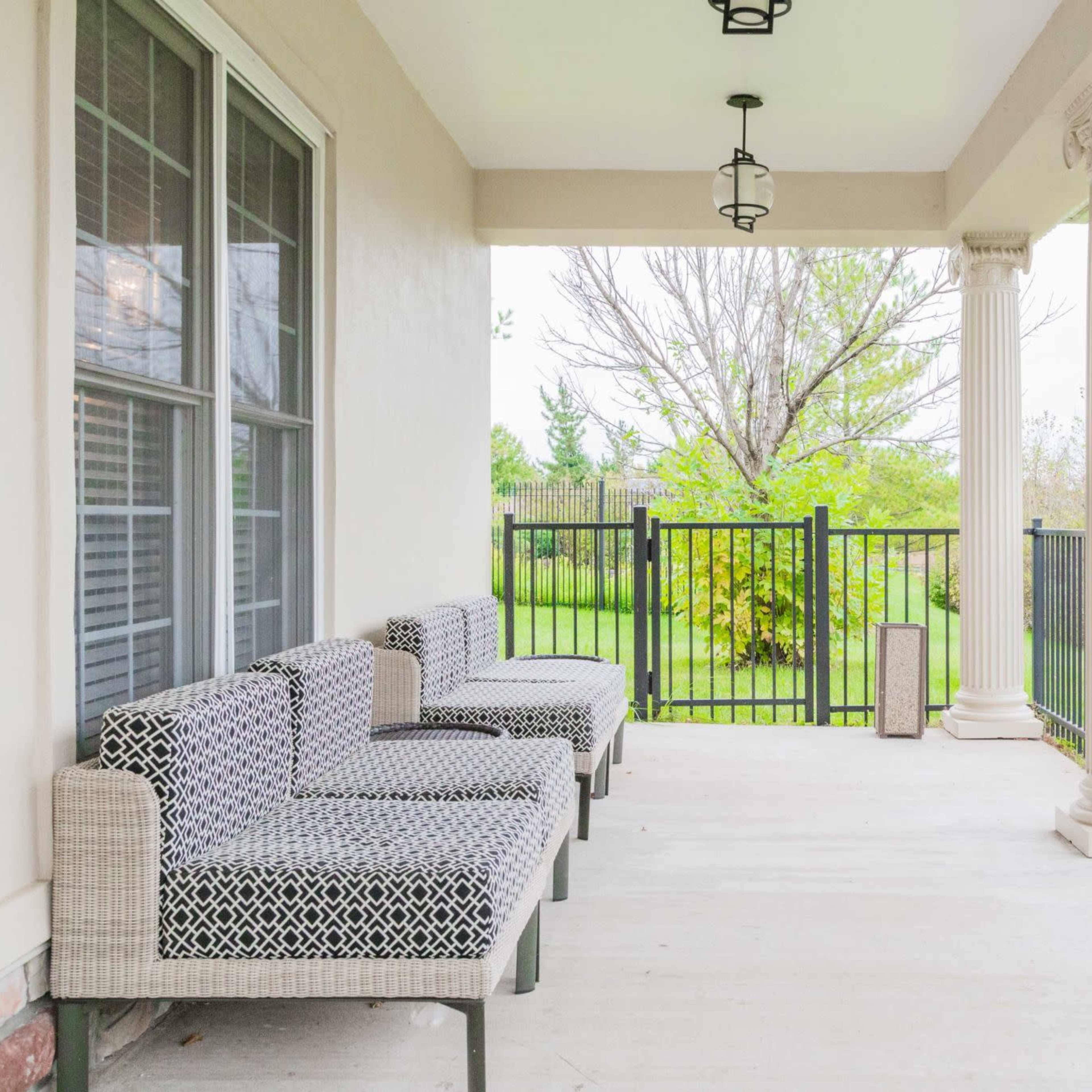 A porch features patterned seating along one side, with decorative columns and a view of a green yard beyond a black metal fence.