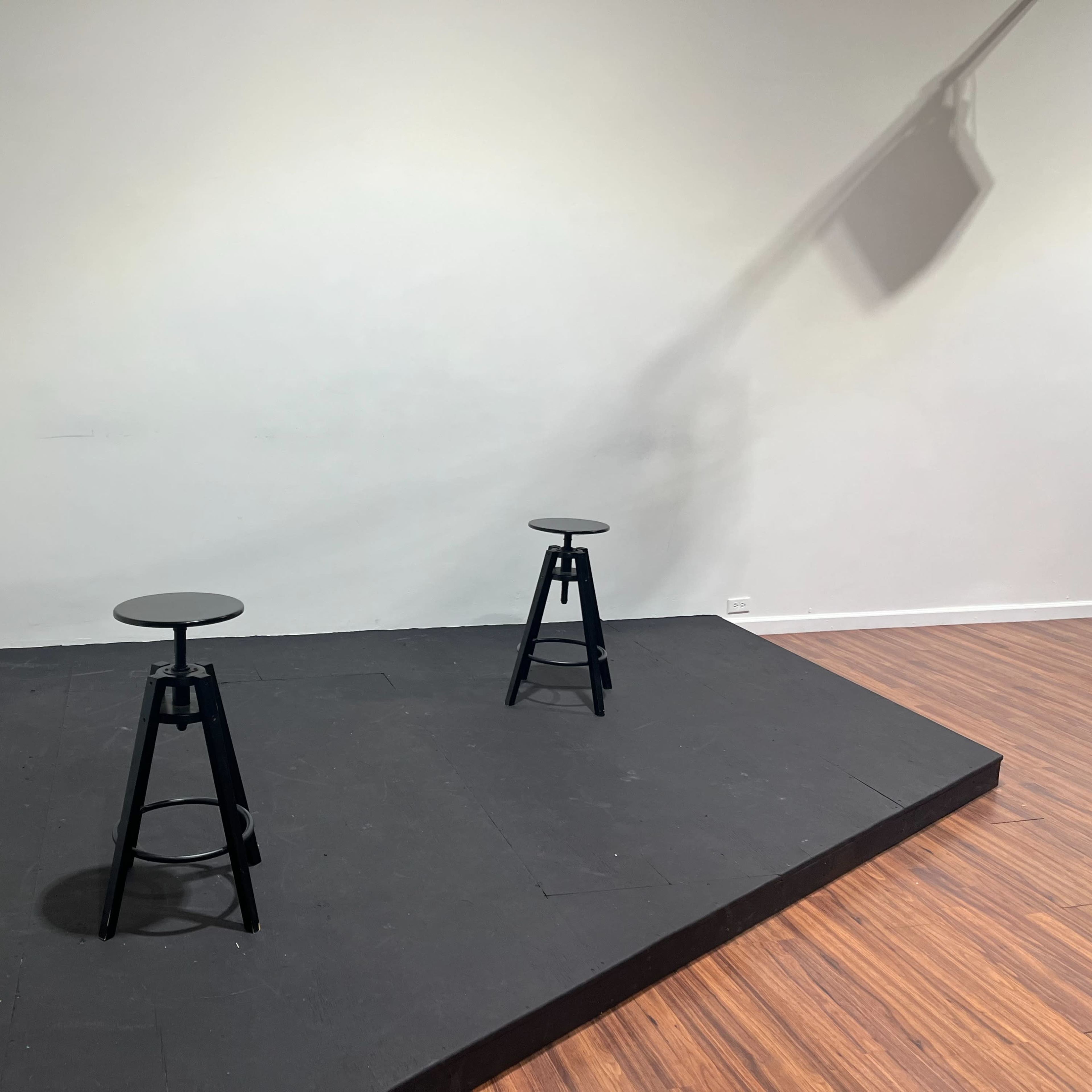 Two black stools positioned on a raised, black stage with a light casting a shadow on the wall.