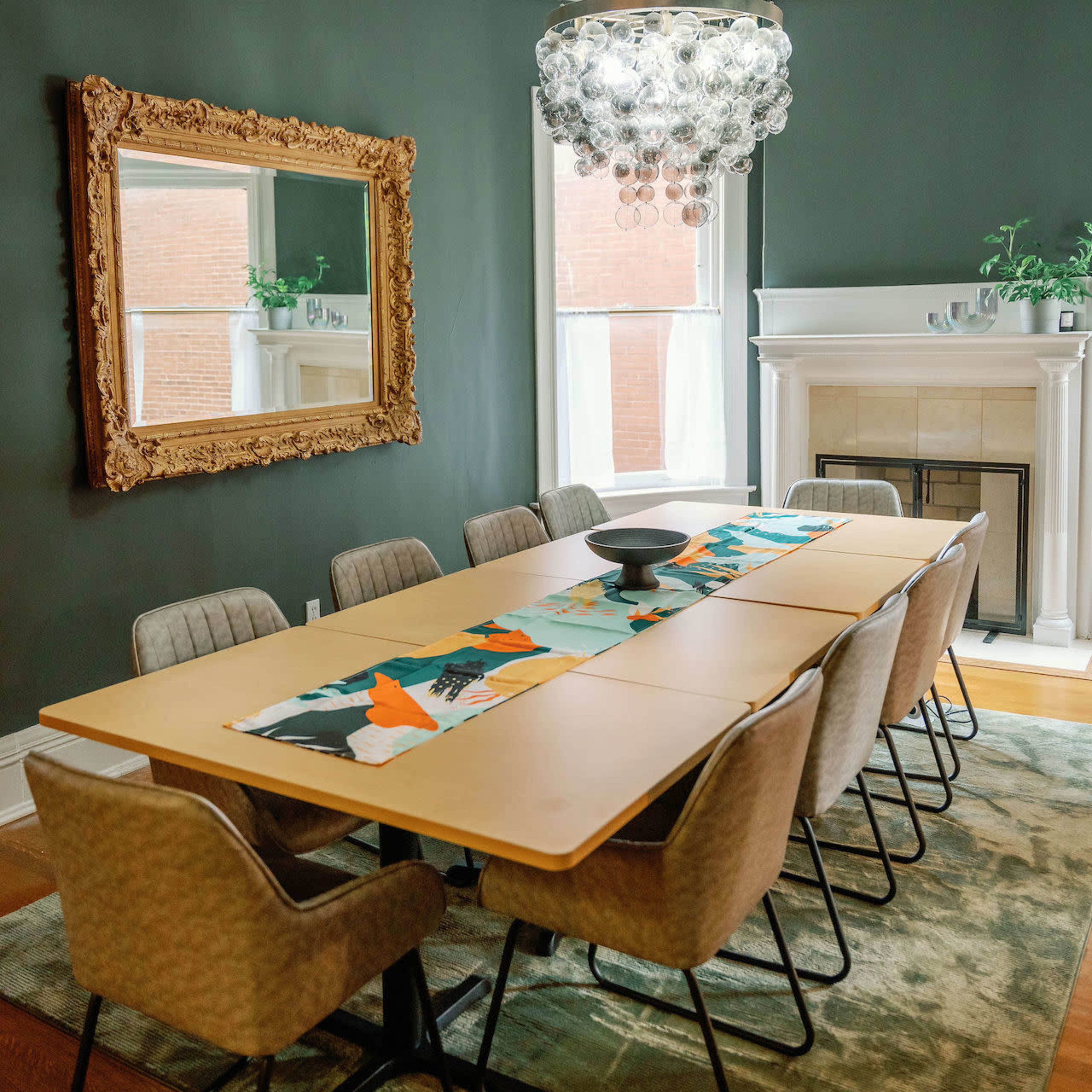A large dining table is set in a stylish room featuring a decorative mirror, an elegant chandelier, and a fireplace.
