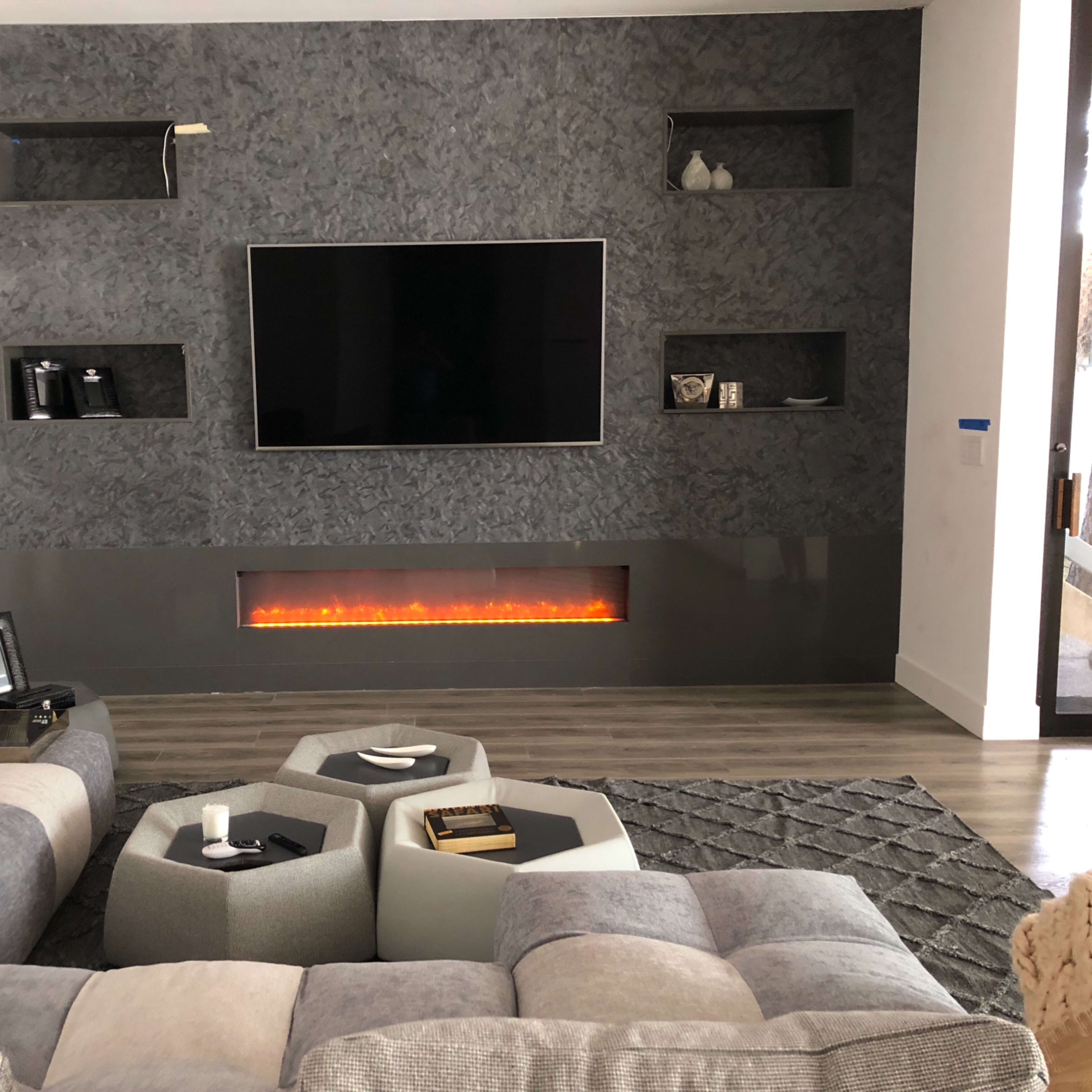 A modern living room with a large television mounted on a textured wall, flanked by minimalist shelves and an electric fireplace below, surrounded by a sectional sofa and geometric coffee tables.