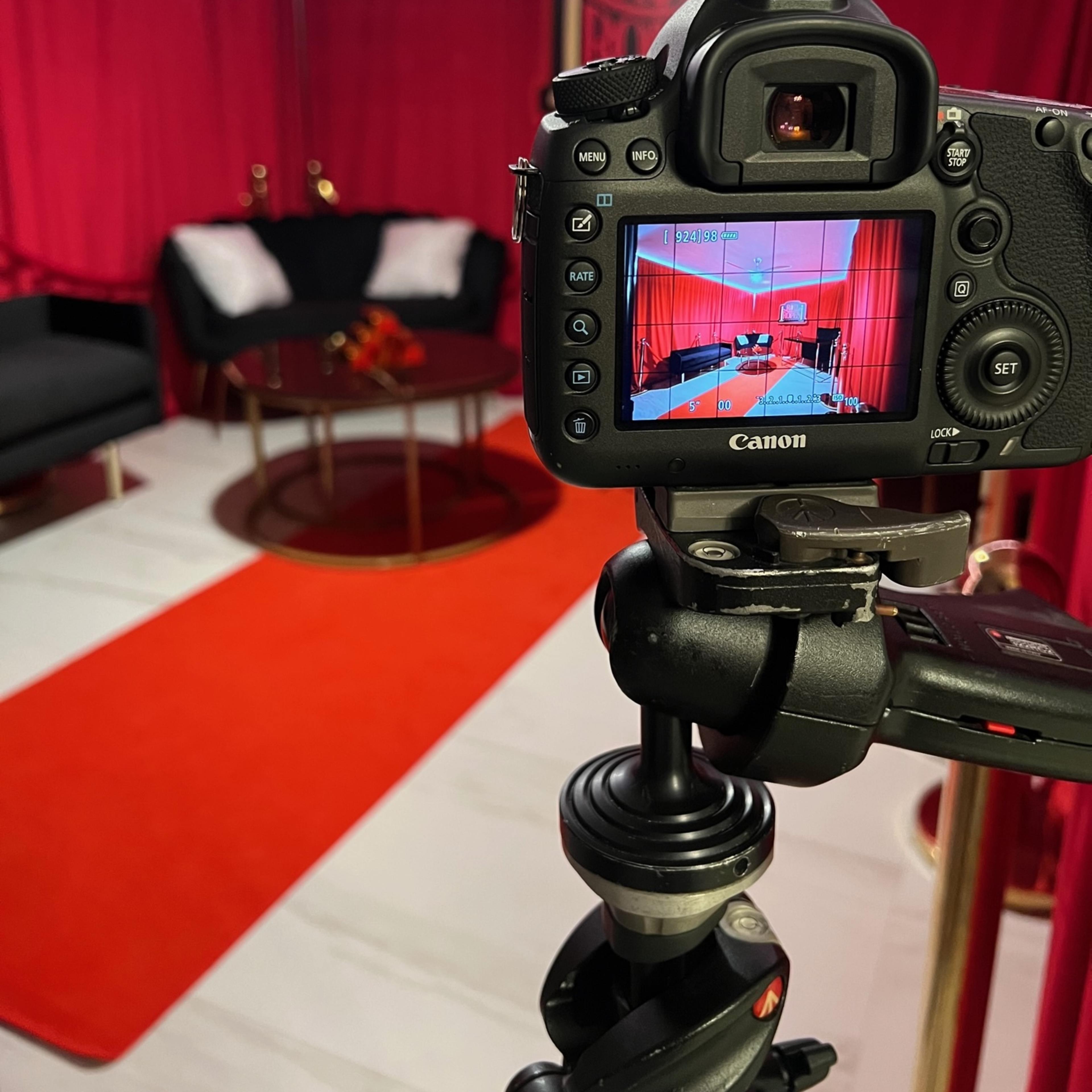 A camera on a tripod captures a red-themed set featuring a black couch, a round table, and a red carpet.