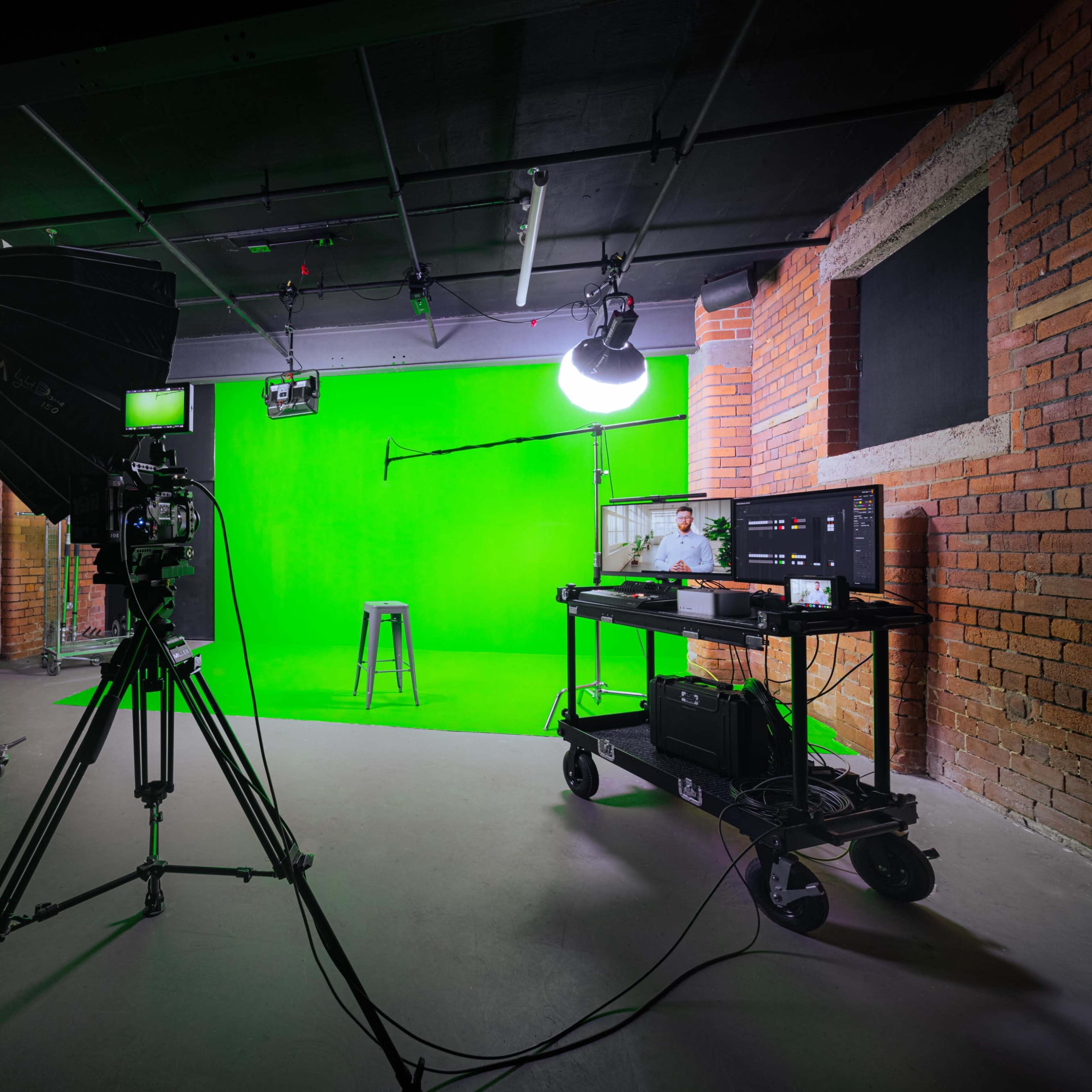 A studio setup featuring a green screen, camera equipment, lighting, and a stool in a brick-walled room.