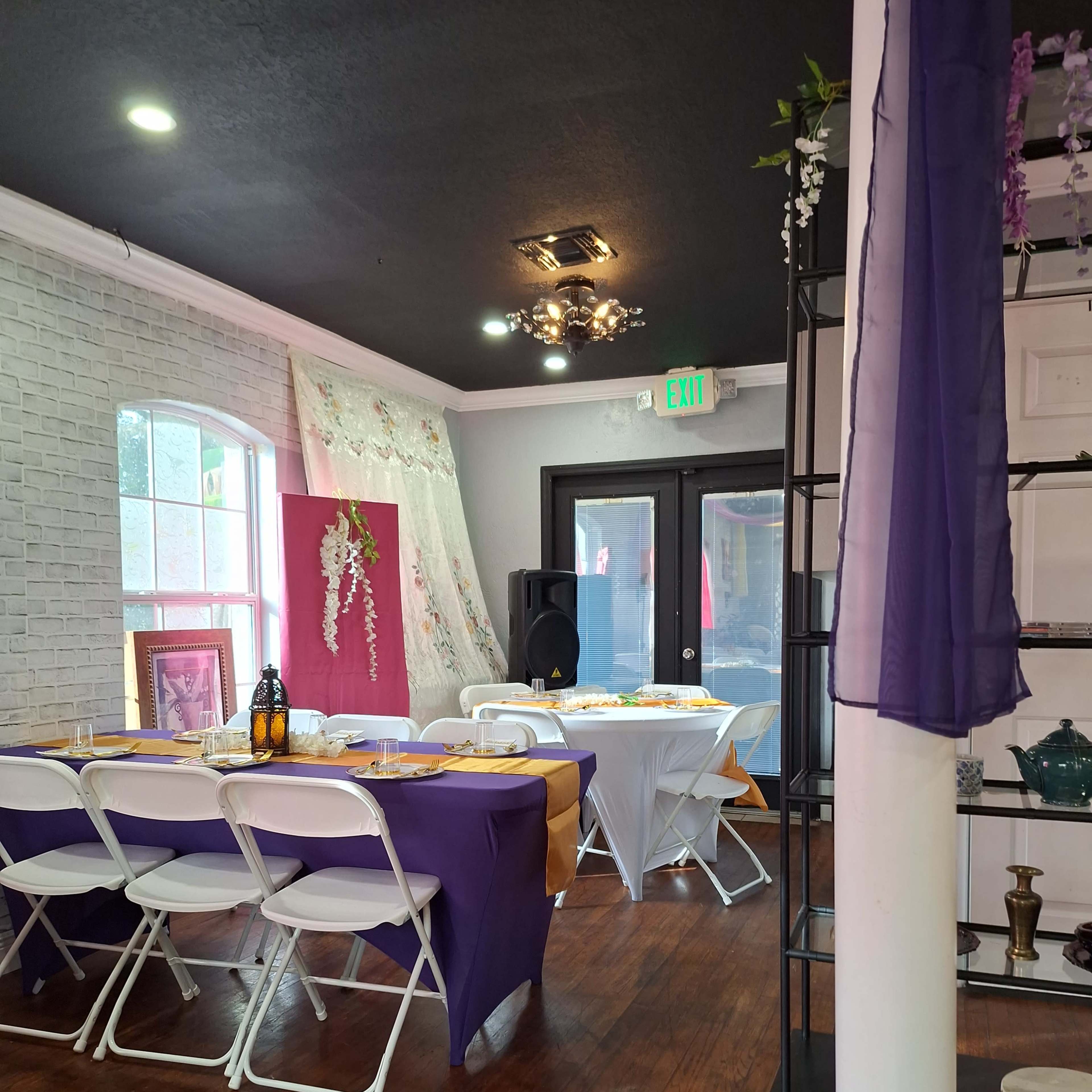 An elegantly decorated event space featuring tables set with purple and white tablecloths, a speaker system, and decorative elements against a brick wall.
