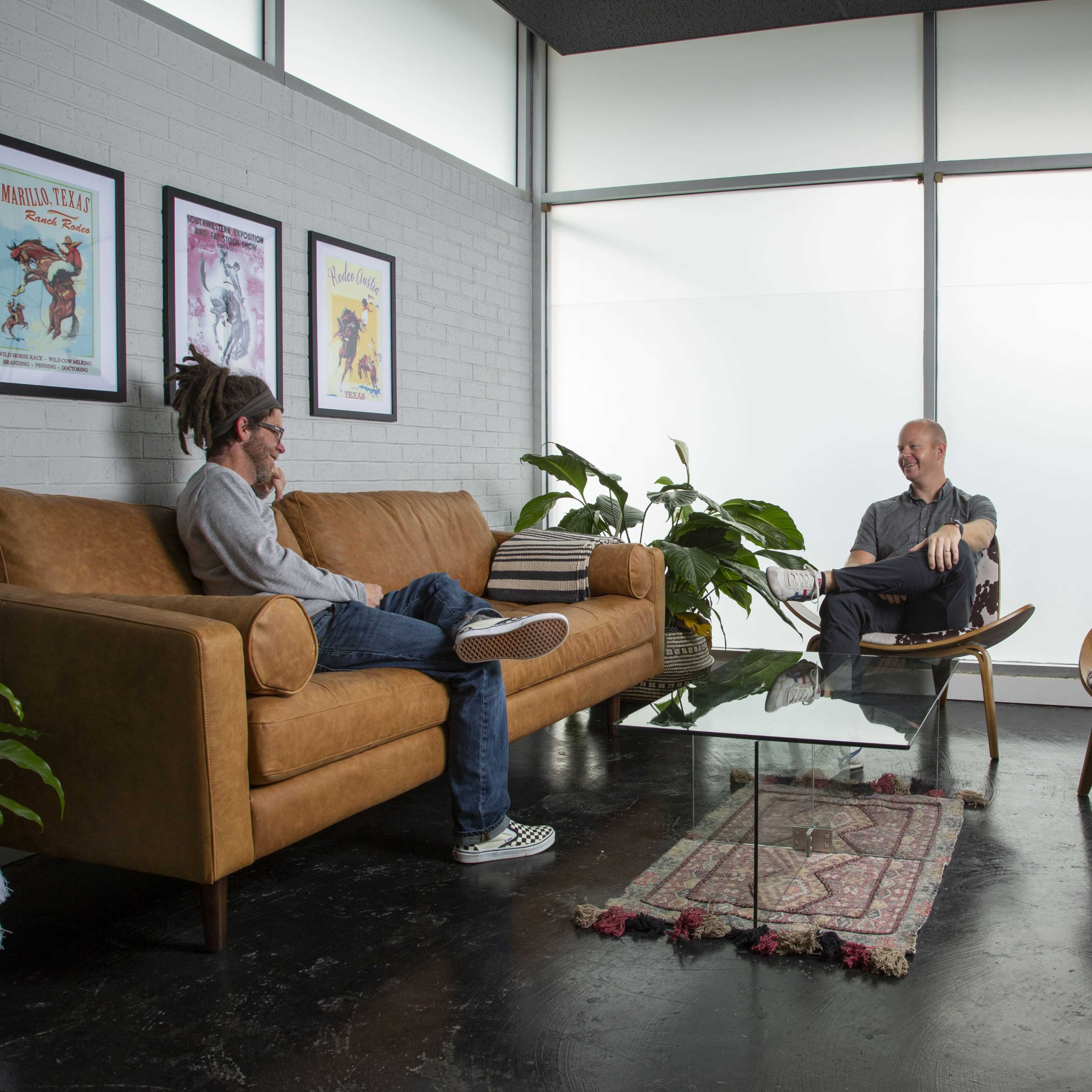 Two men are seated in a modern room, one on a brown leather sofa and the other in a chair, with plants and a glass coffee table between them.