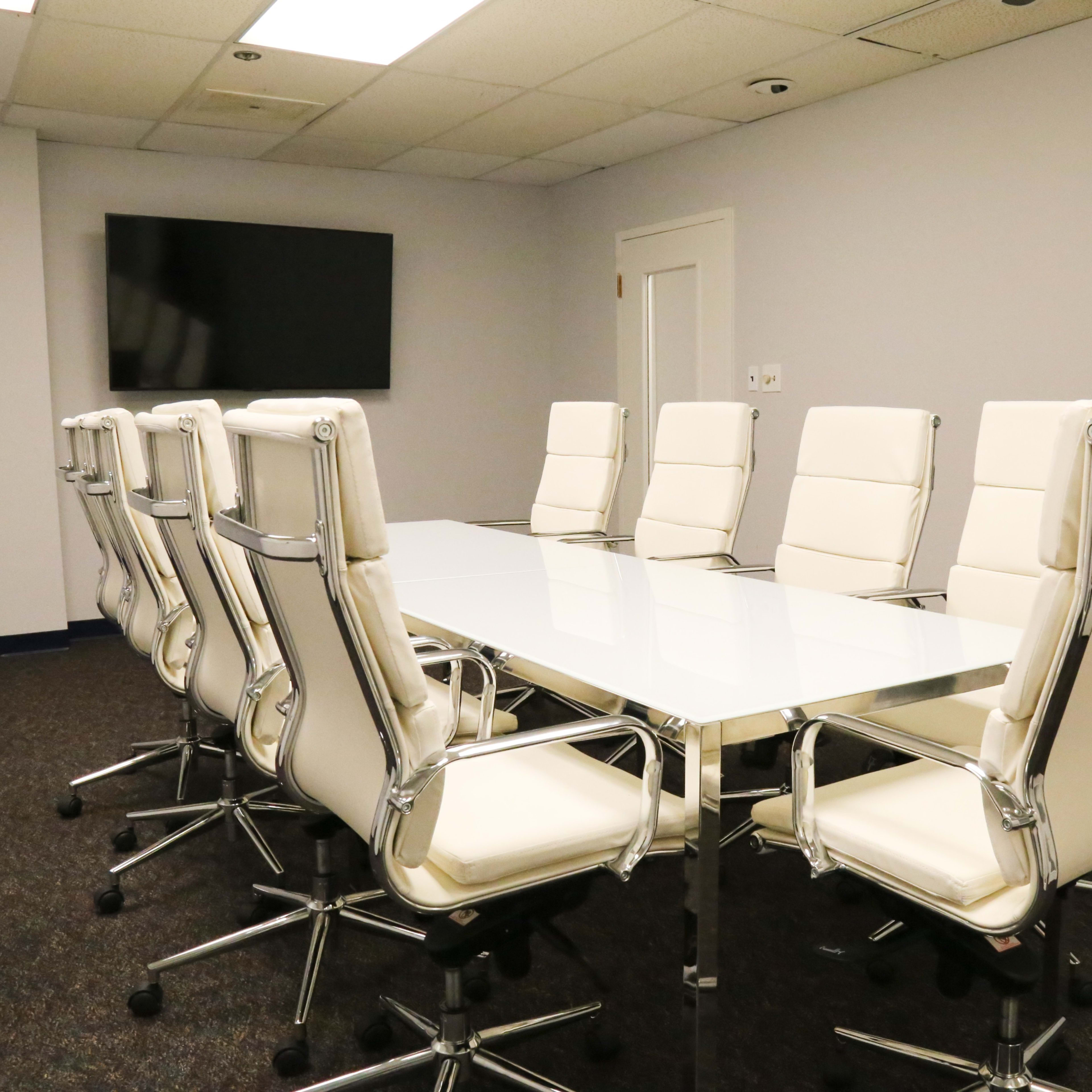 A modern conference room features a long glass table with white leather chairs arranged around it and a television mounted on the wall.