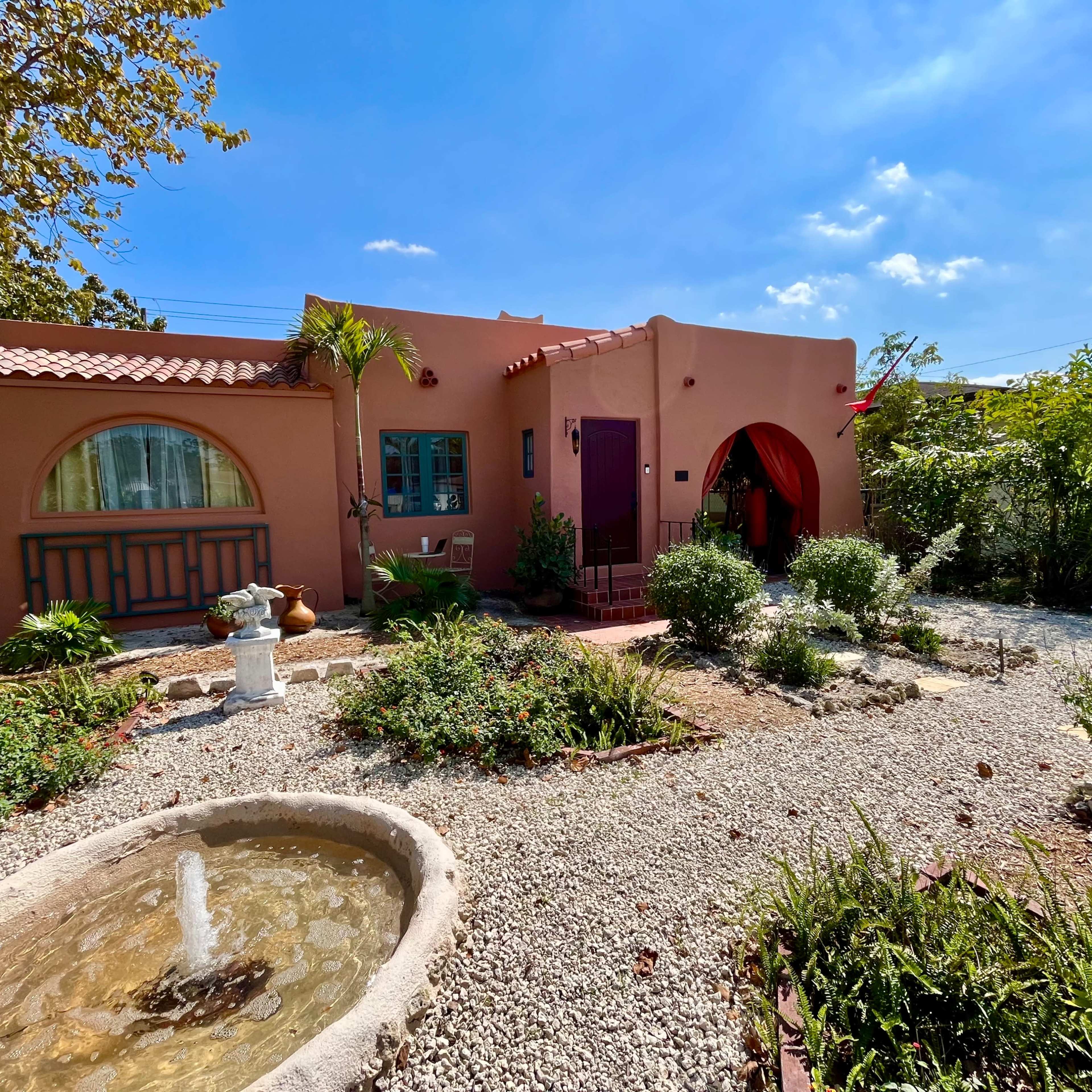 A pink stucco house with arched windows and a red door is surrounded by a garden featuring a stone fountain and various plants.
