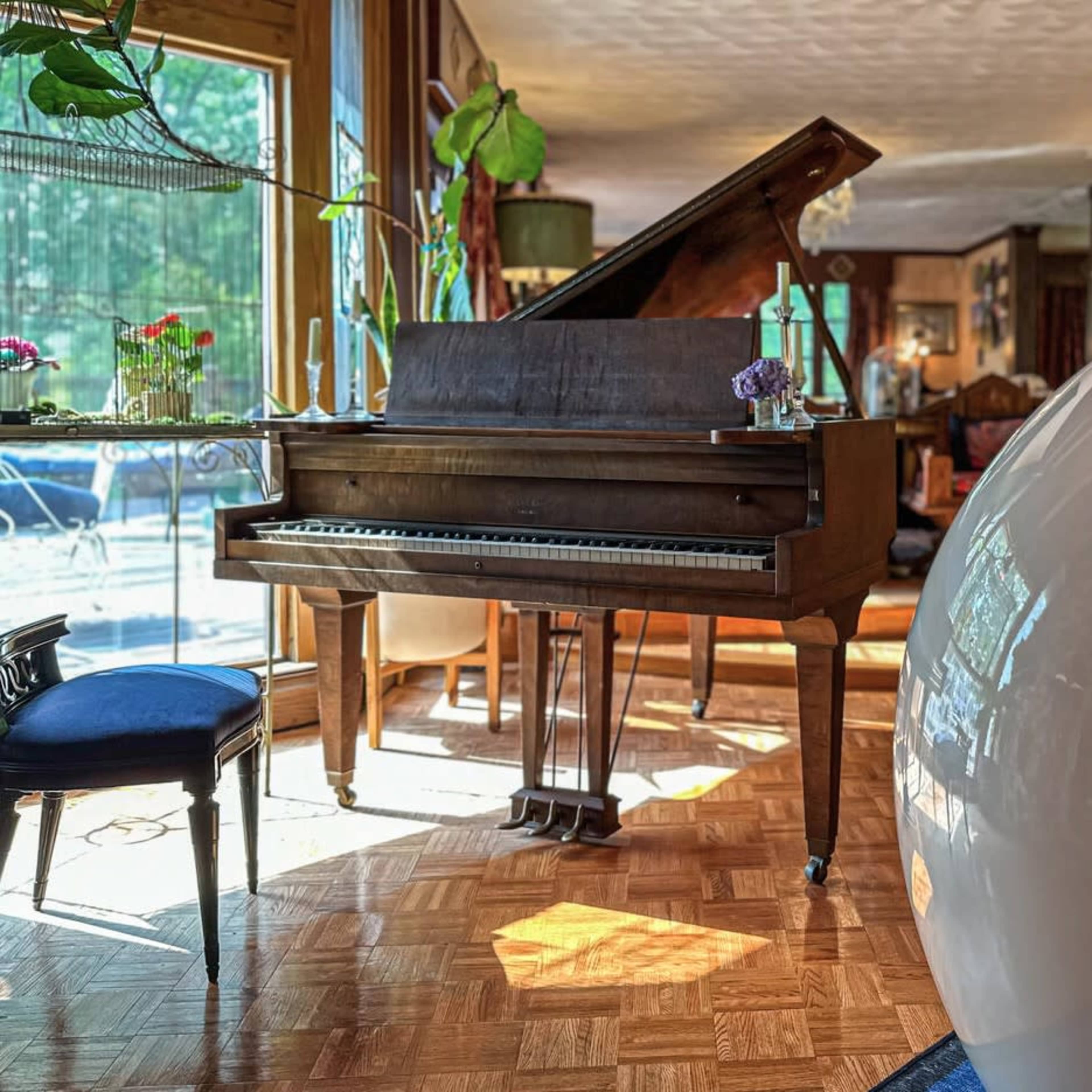 A wooden grand piano beside a curved white lounge chair in a room with large windows and various decorative elements.