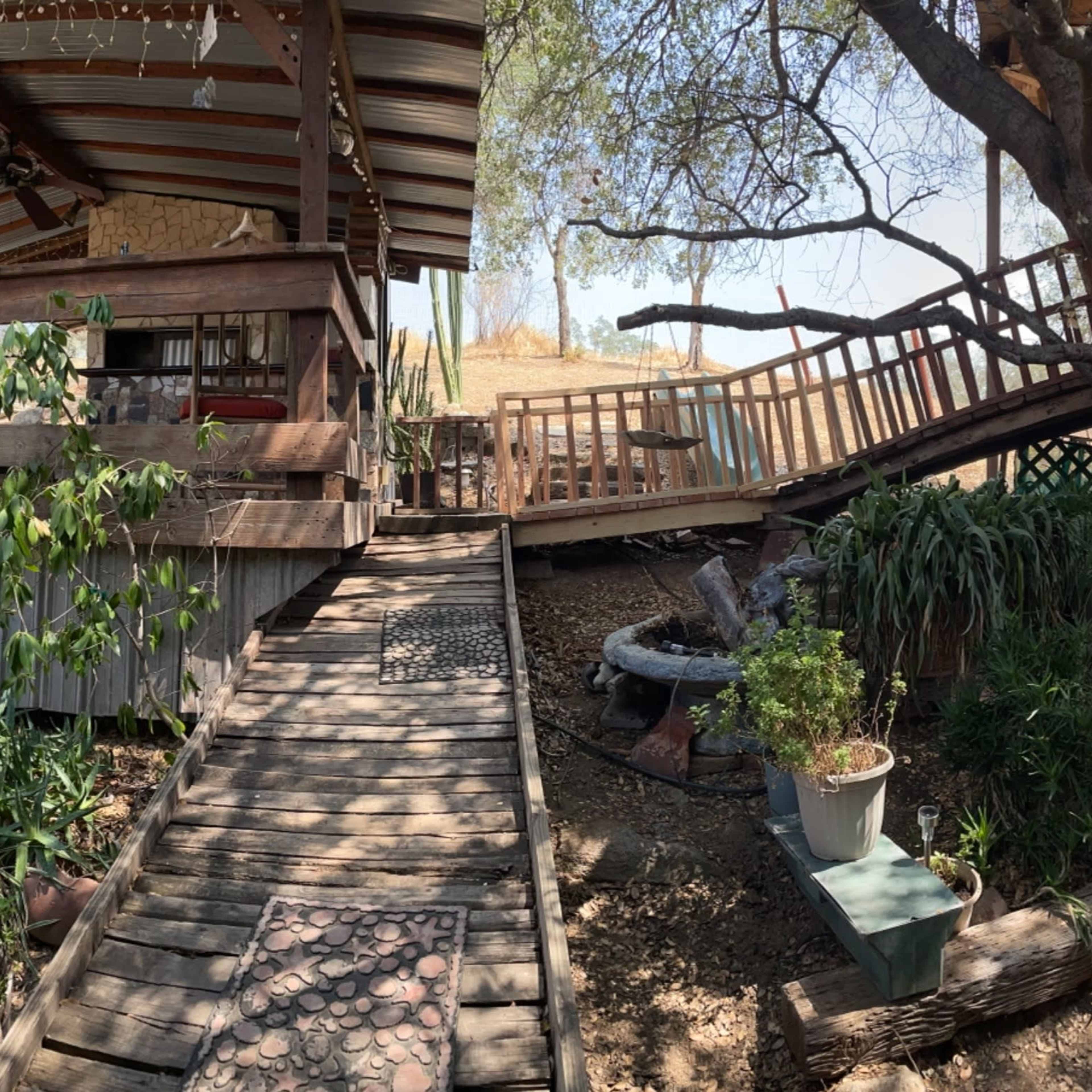 A wooden deck and garden area with various plants, leading to a house elevated on stilts under a sunny sky.