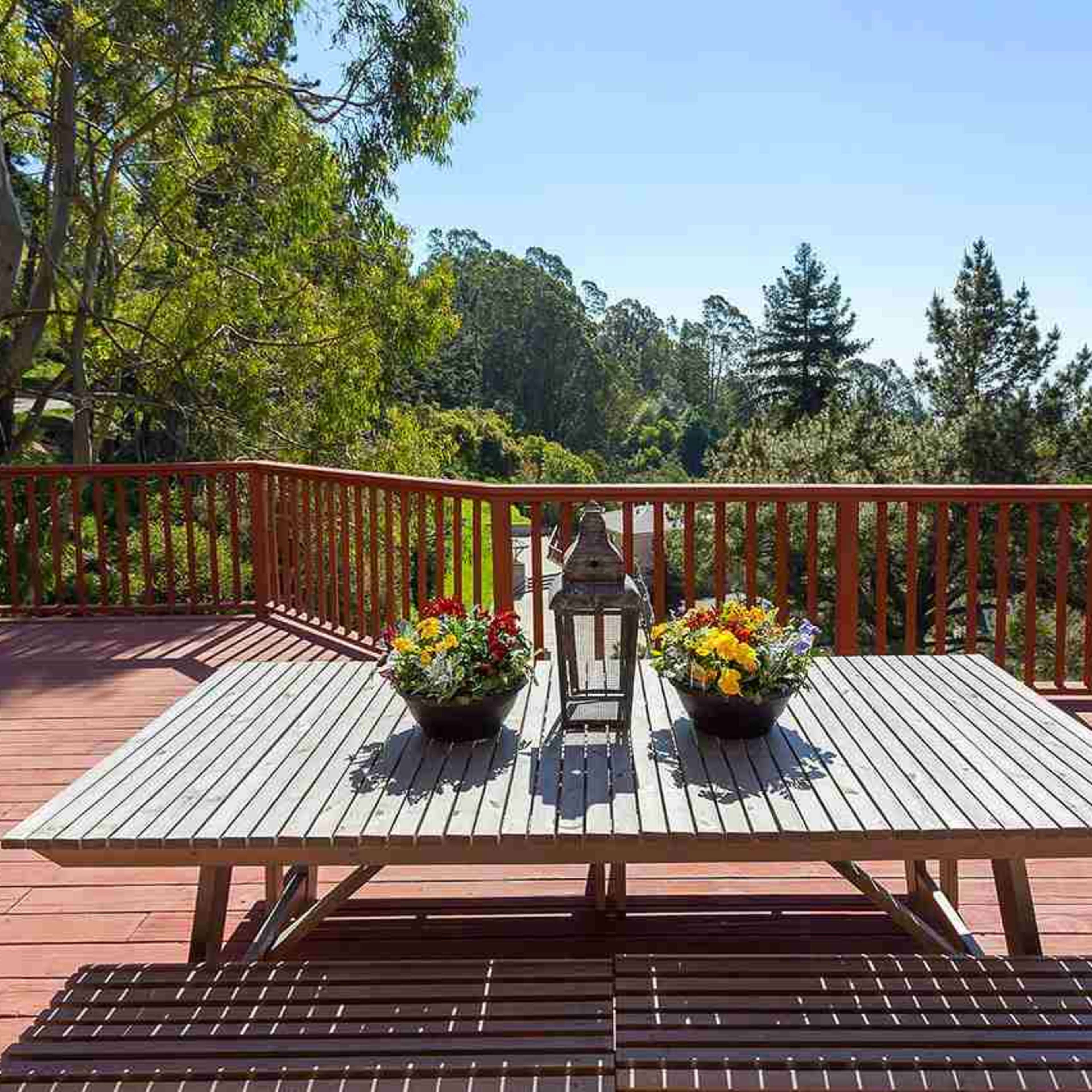 A wooden deck features a table adorned with flower pots and a lantern, overlooking a lush green landscape.