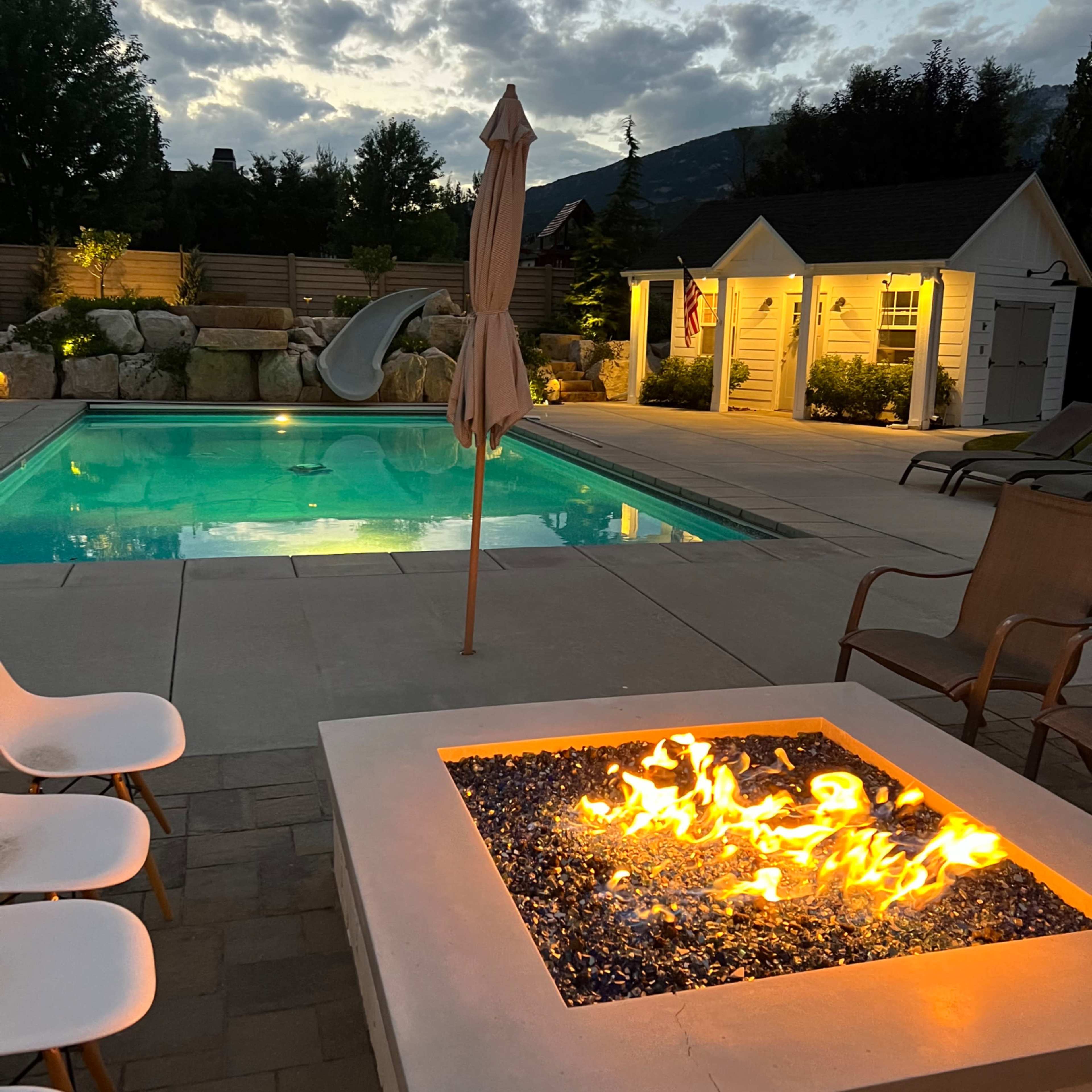 A backyard pool area featuring a lit fire pit in the foreground, a pool with a water slide in the background, and a partially cloudy sky above.