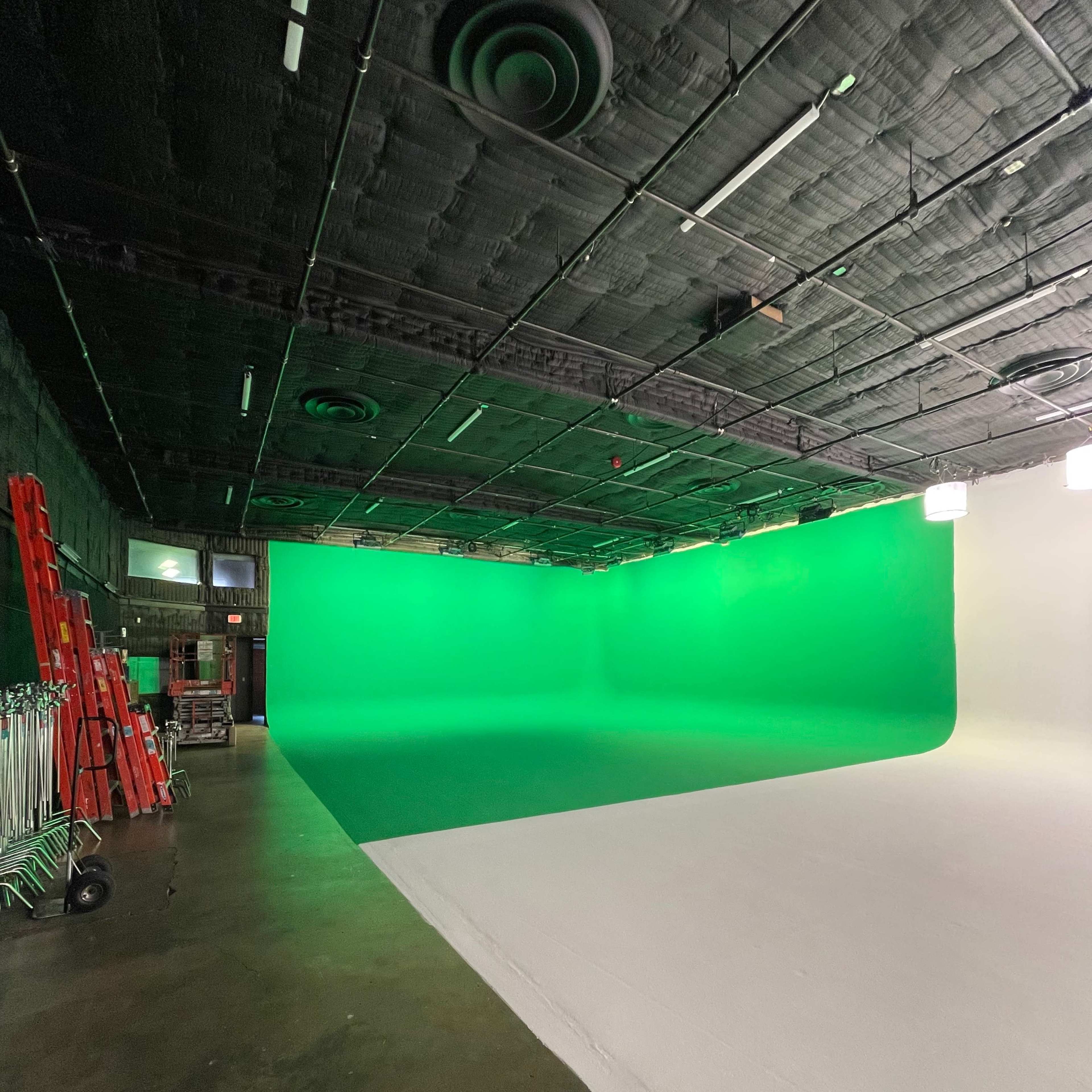 A studio with a green screen on one side and a white surface on the other, surrounded by lighting equipment and ladders.