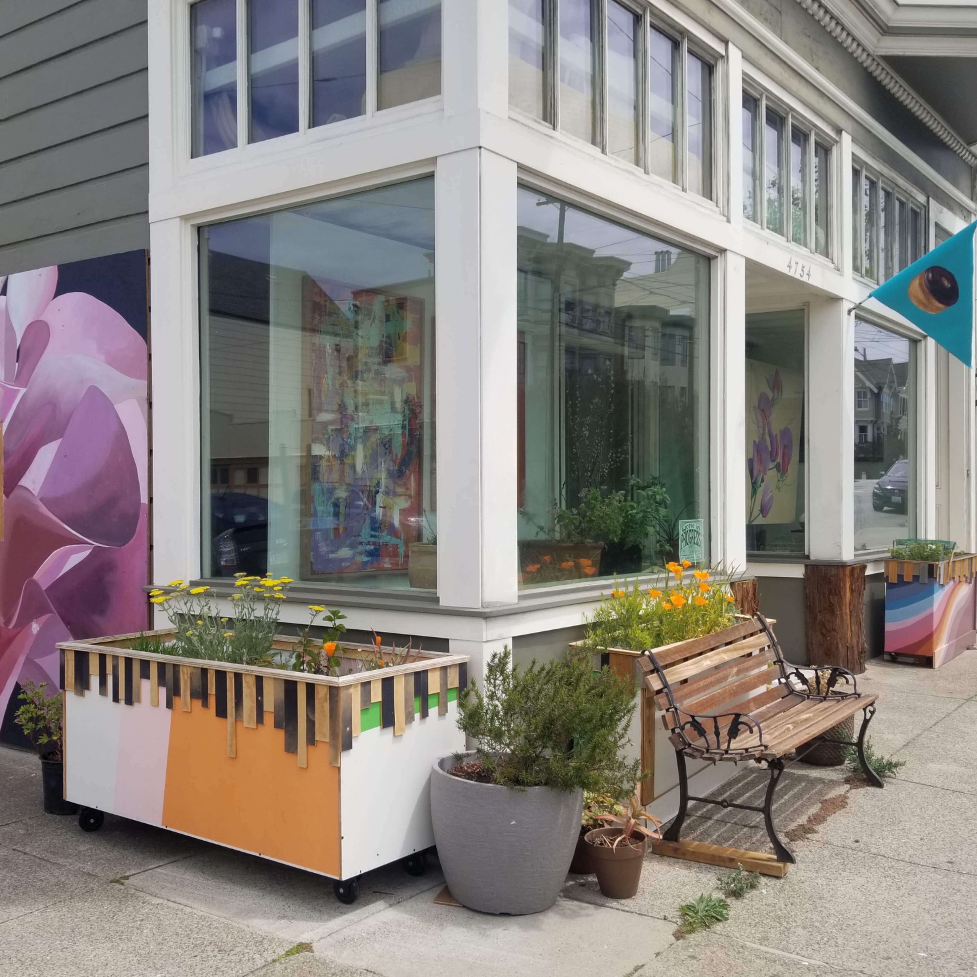 A colorful mural of a pink flower decorates the side of a building, which features large windows, planters with flowers, and a bench in front.