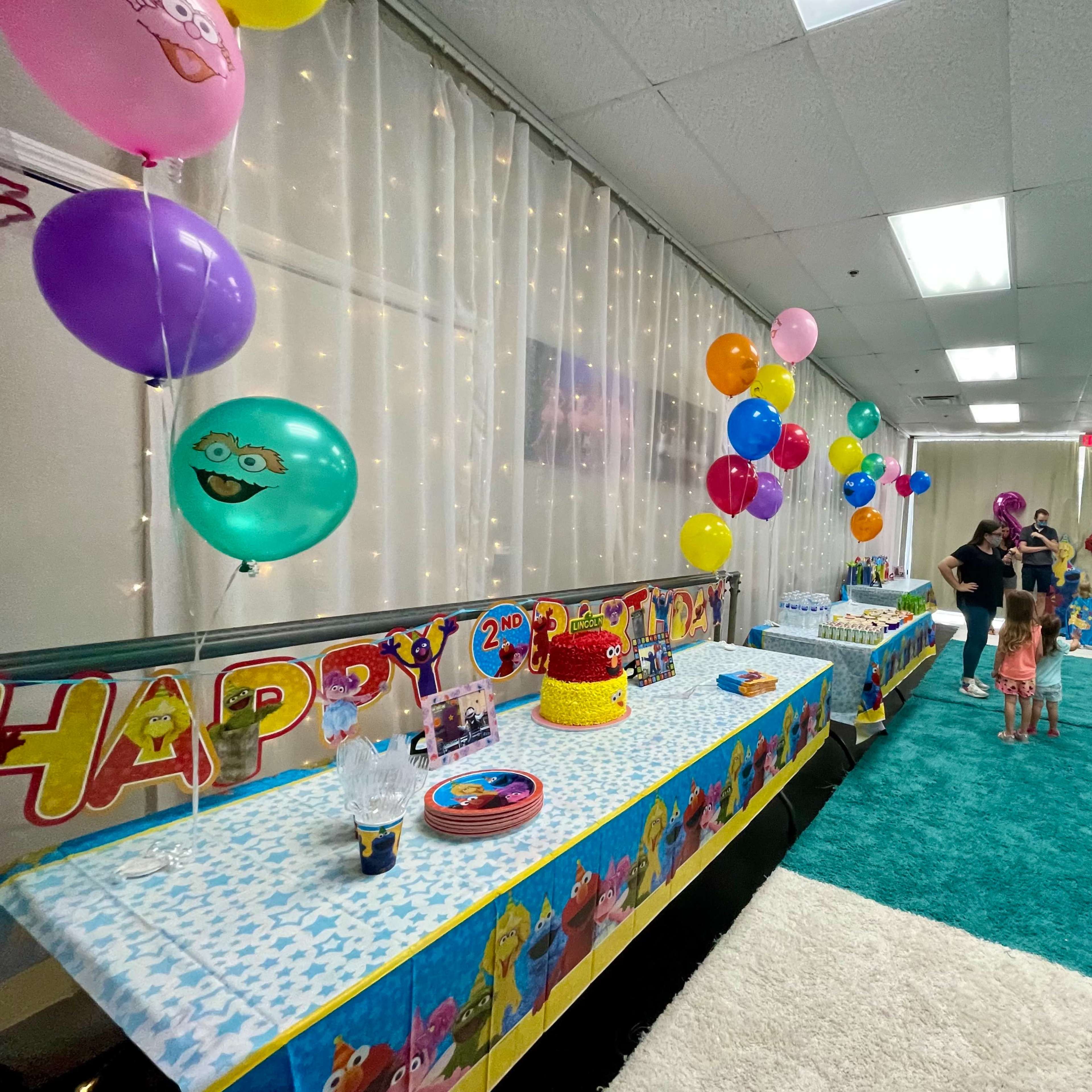 A colorful children's birthday party setup features decorated tables, balloons, and a cake, with guests mingling in a brightly lit room.