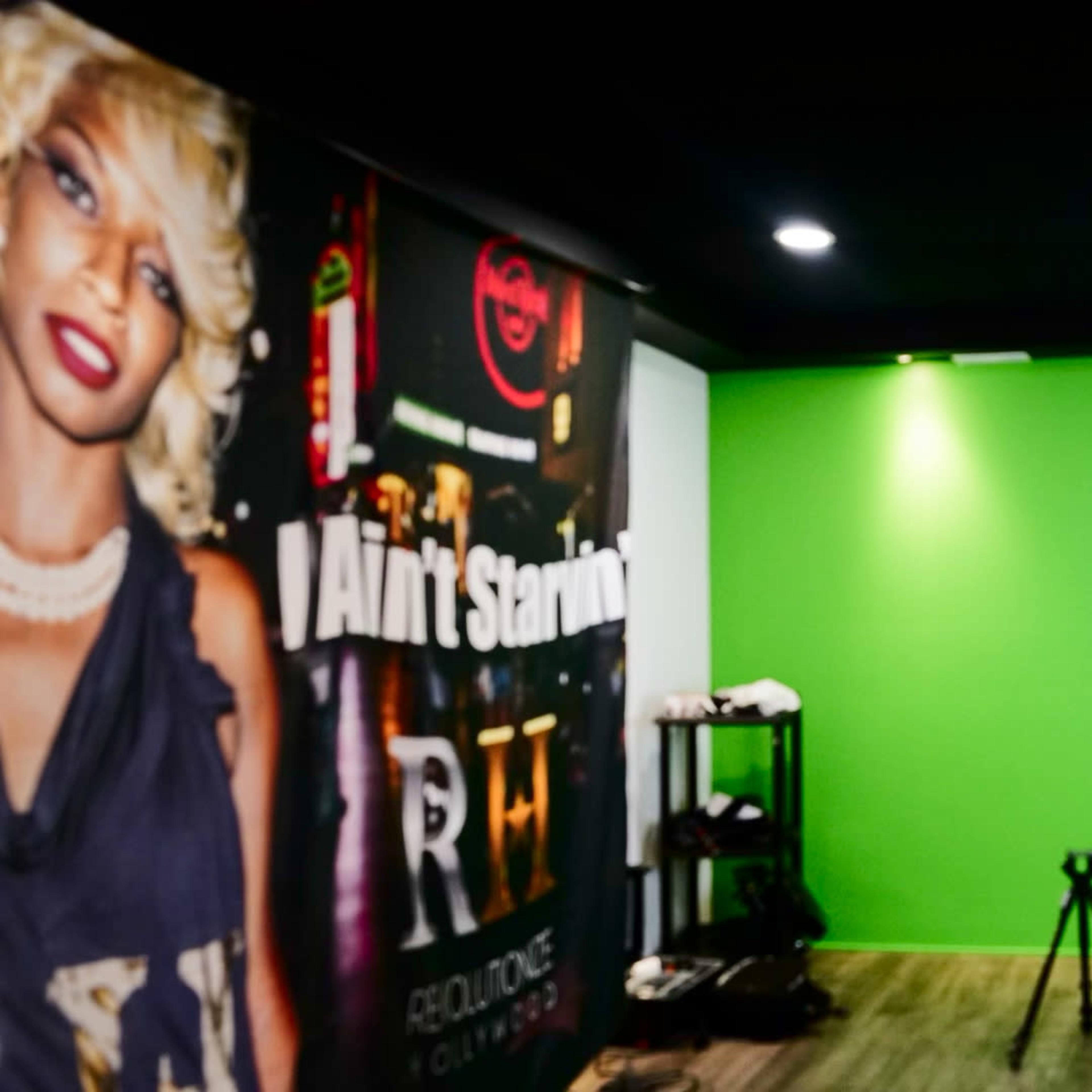 An empty studio space featuring a green backdrop, a camera tripod, and lighting equipment against a wall with a large mural.