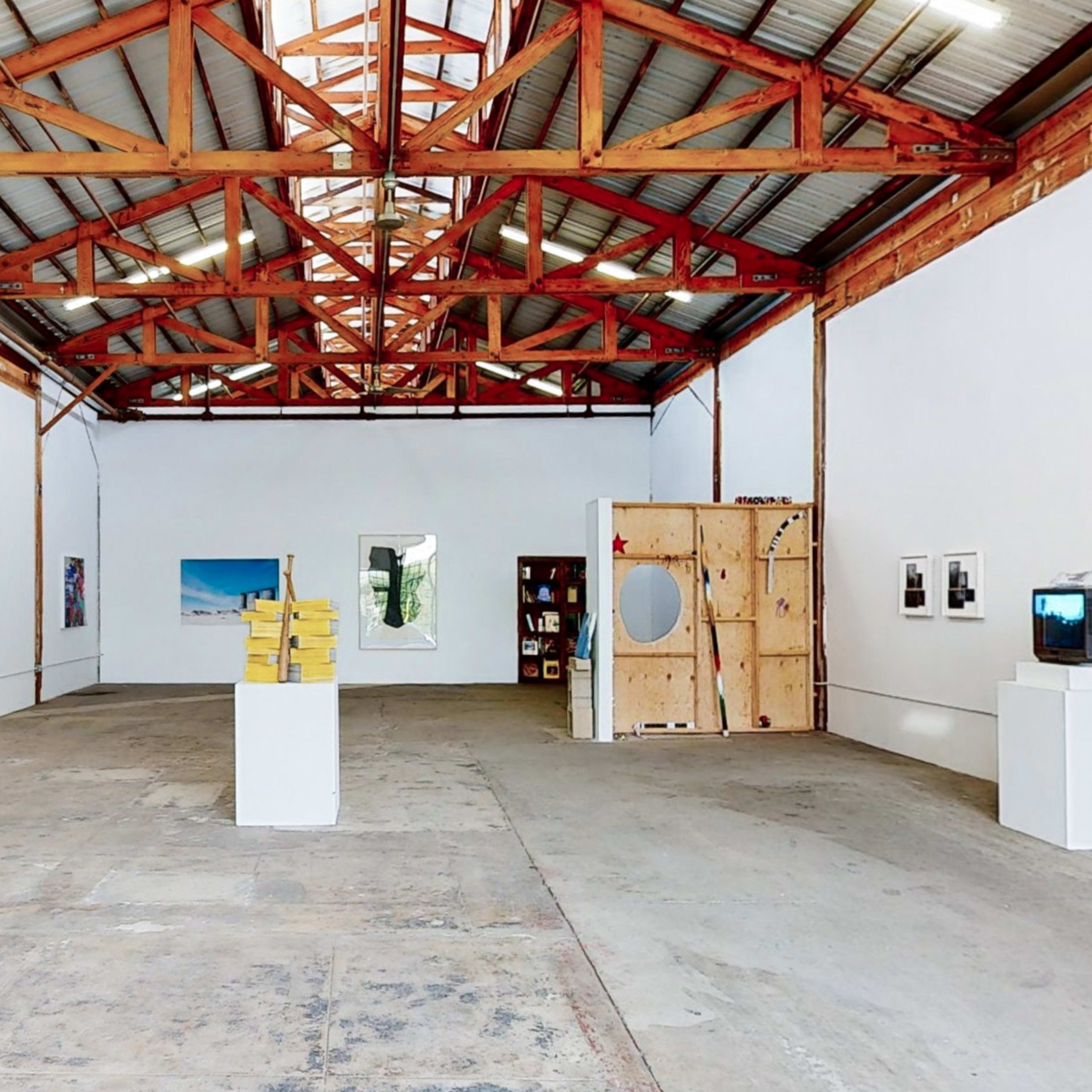 An art gallery with a high ceiling, featuring various artworks displayed on white walls and a wooden structure in the background.